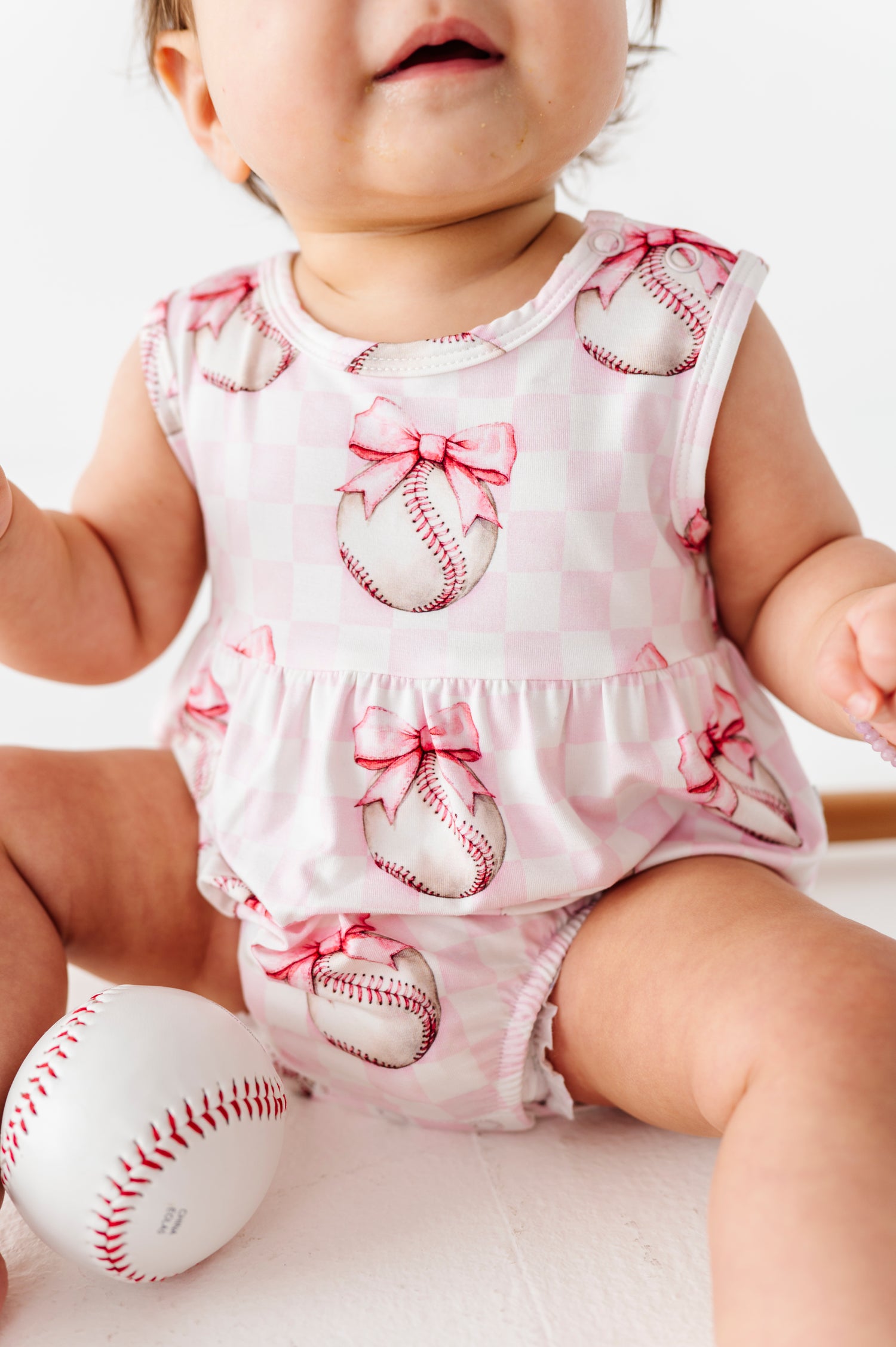 Baby wearing a pink bamboo romper with baseball and bow designs, sitting on a white surface.