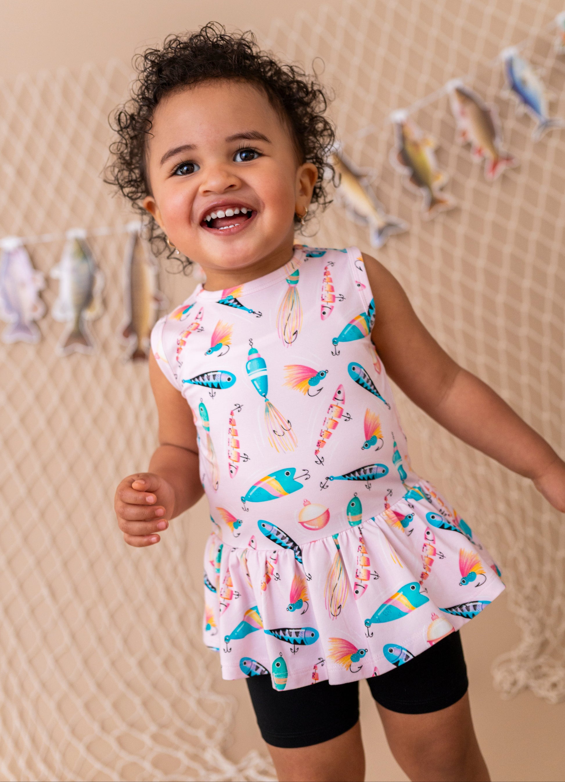Child wearing a colorful dress with a pattern of fishing lures, standing against a beige wall with decorative elements.