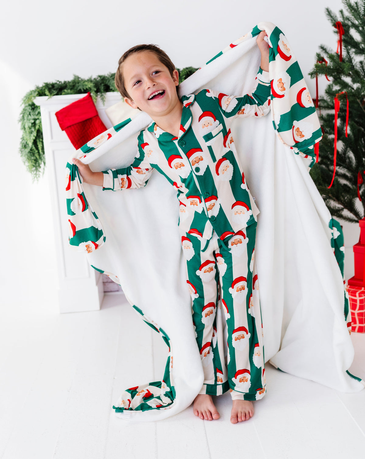 A young child wearing festive Christmas pajamas with a Santa Claus pattern, playfully holding up a large white blanket in a bright, holiday-themed room next to a decorated Christmas tree.