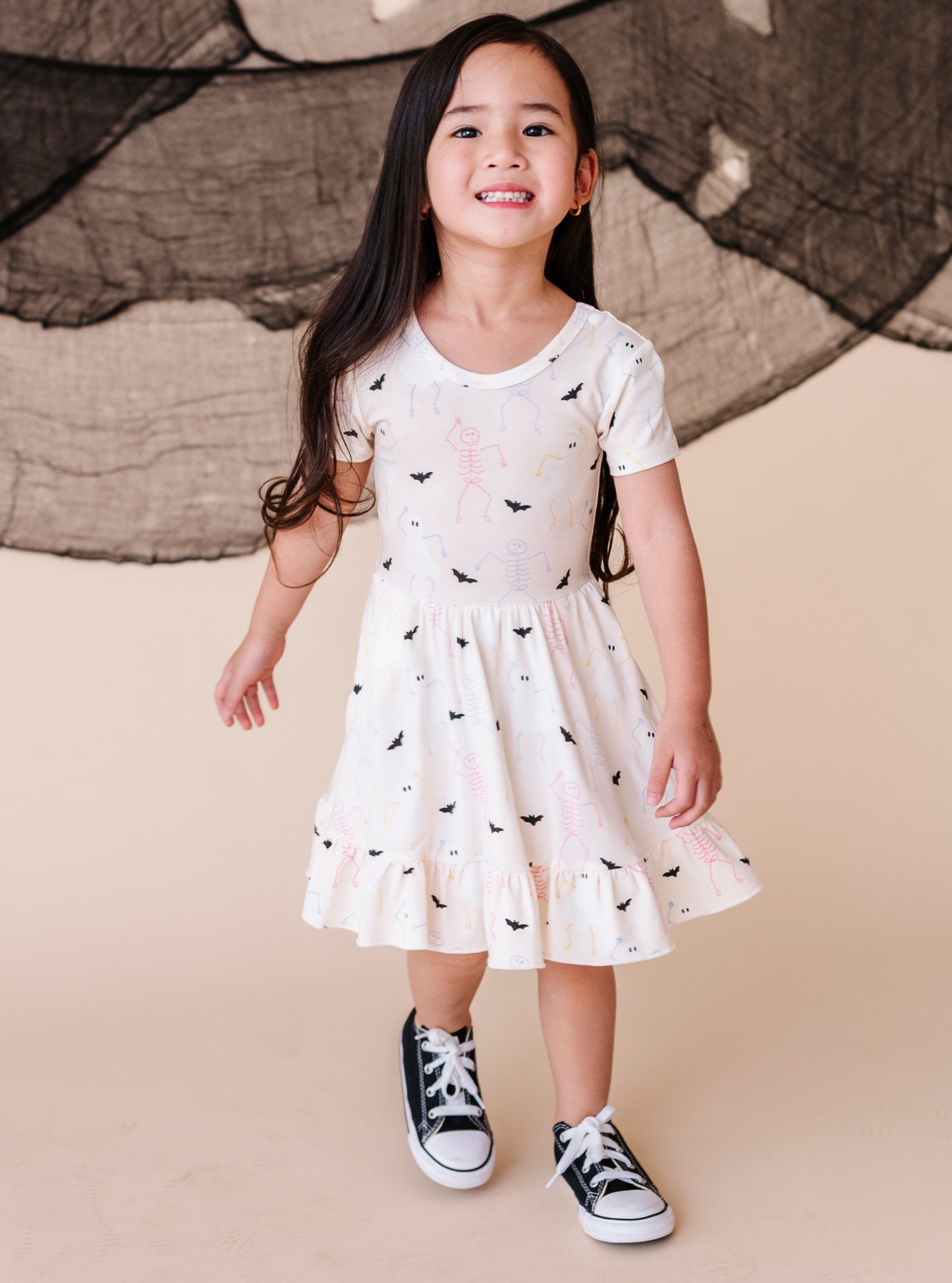 Young girl wearing a cream colored  dress with halloween pattern and black sneakers against a beige background