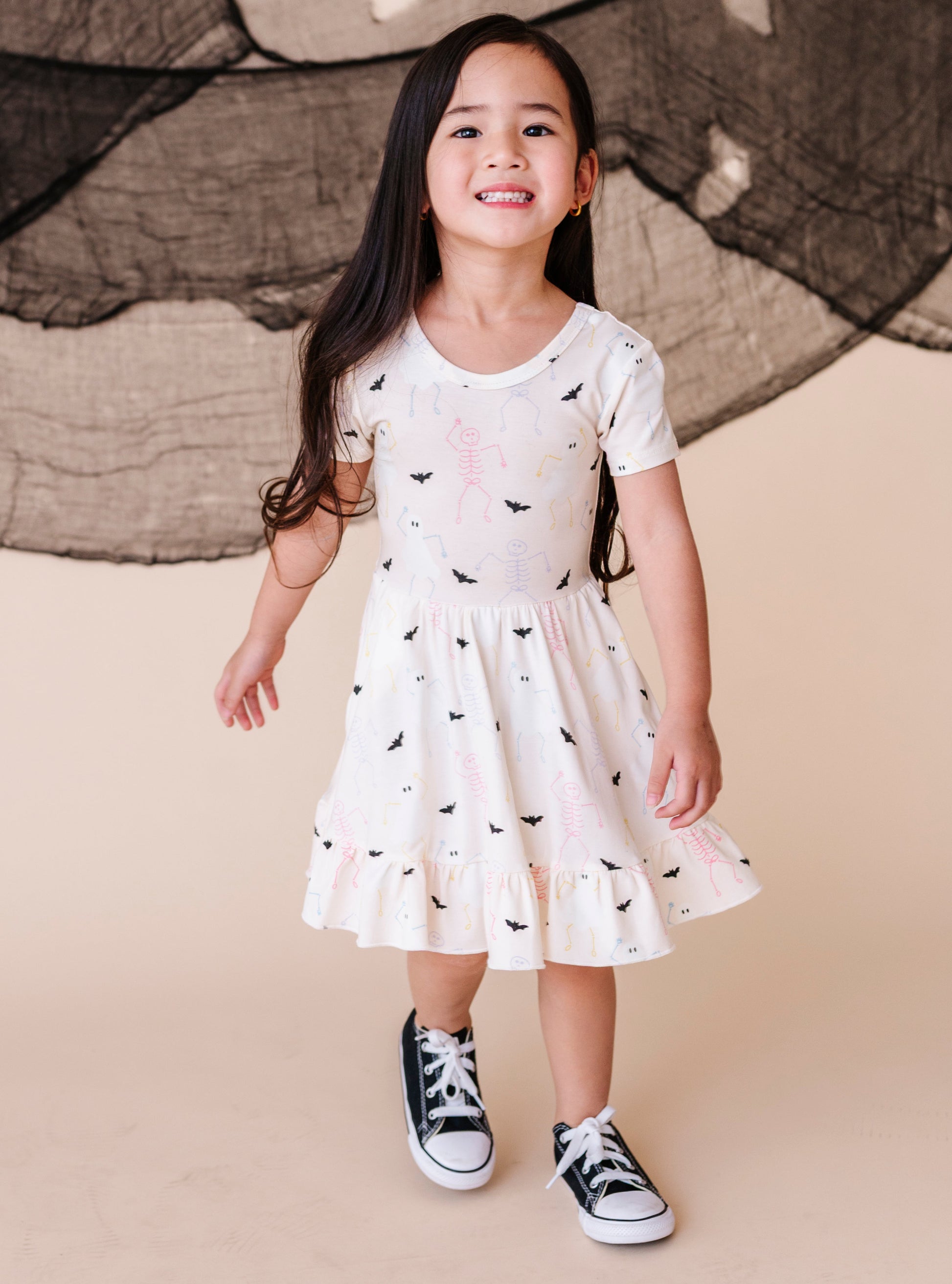 Young girl wearing a cream colored  dress with halloween pattern and black sneakers against a beige background