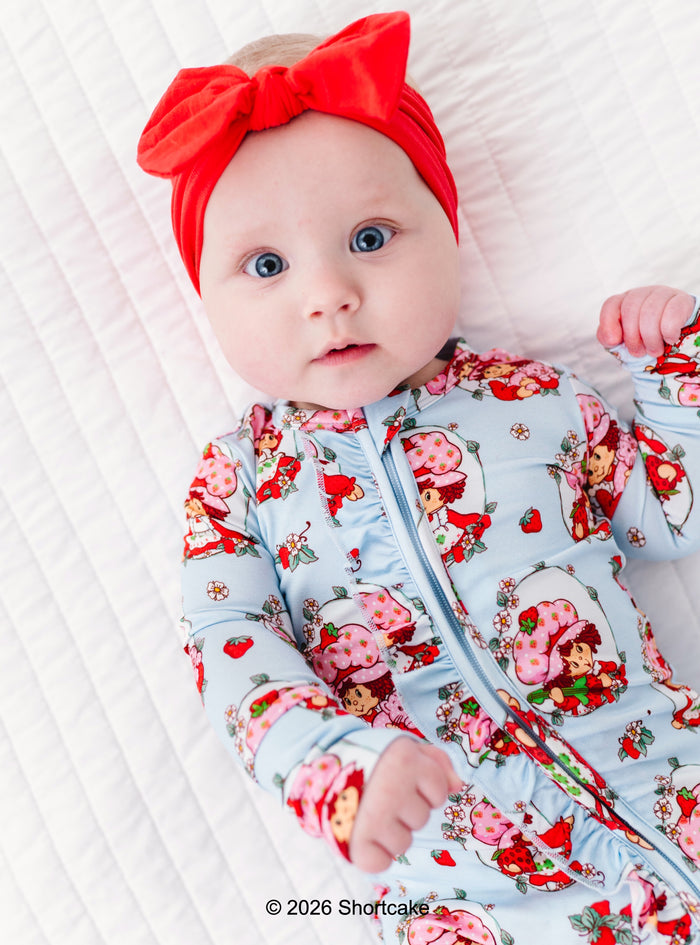 Baby wearing a bamboo Strawberry Shortcake™ convertible romper with a red bow headband on a white background