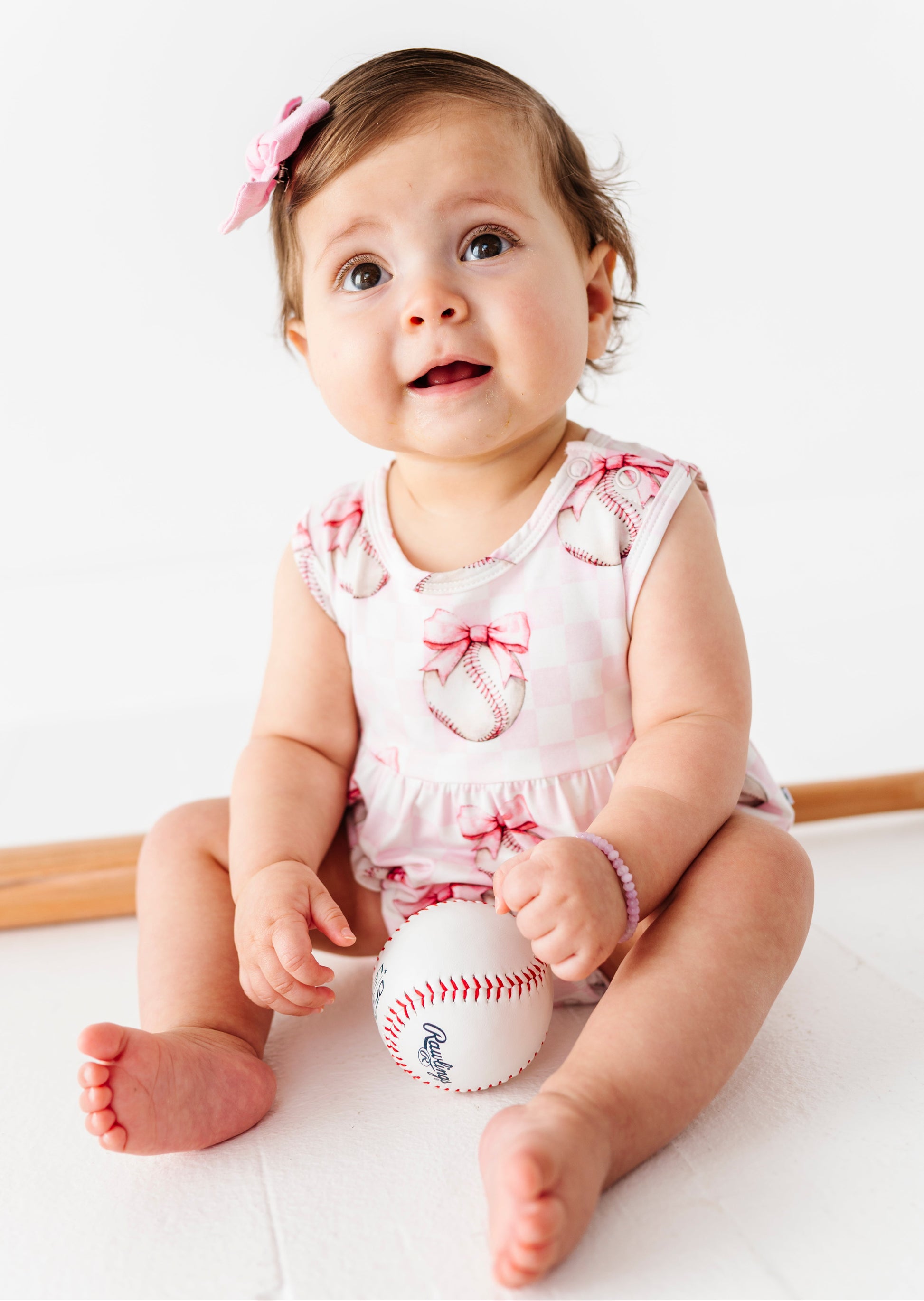 Baby girl sitting on a white surface with a baseball, wearing a pink bamboo baseball outfit.