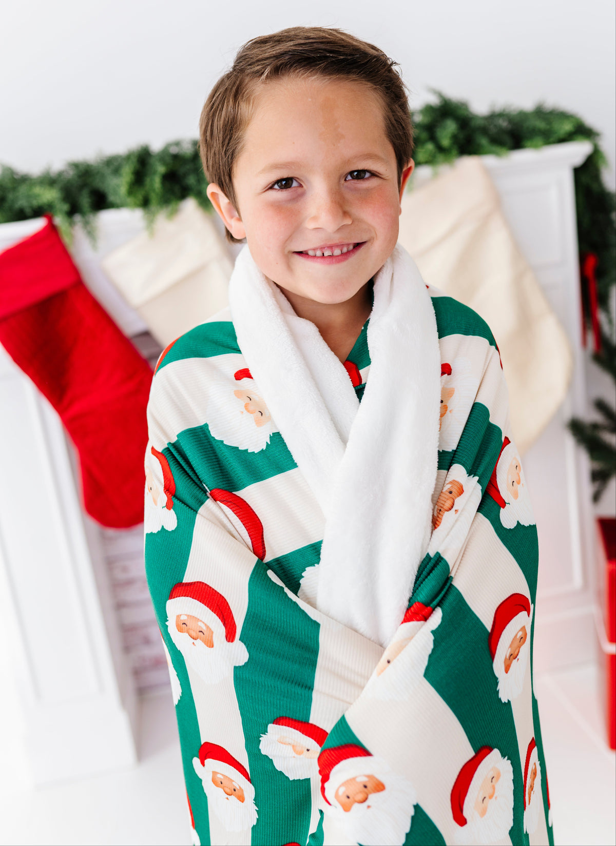 A young boy smiling and wrapped in a festive Christmas-themed bathrobe with a white faux-fur collar. The robe features a green and white pattern of Santa Claus faces.