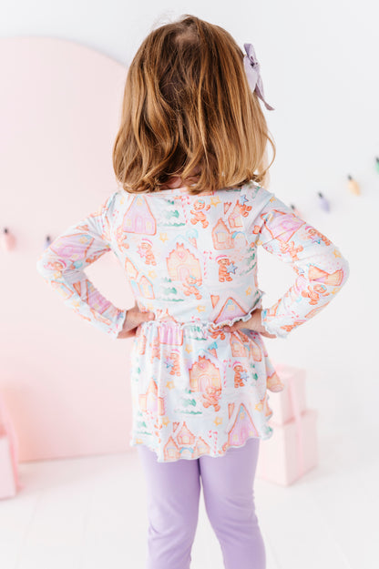 A young girl with blonde hair is shown from behind wearing a long-sleeve bamboo peplum top with a pastel gingerbread village pattern, paired with light purple leggings in a bright, pink-themed room.