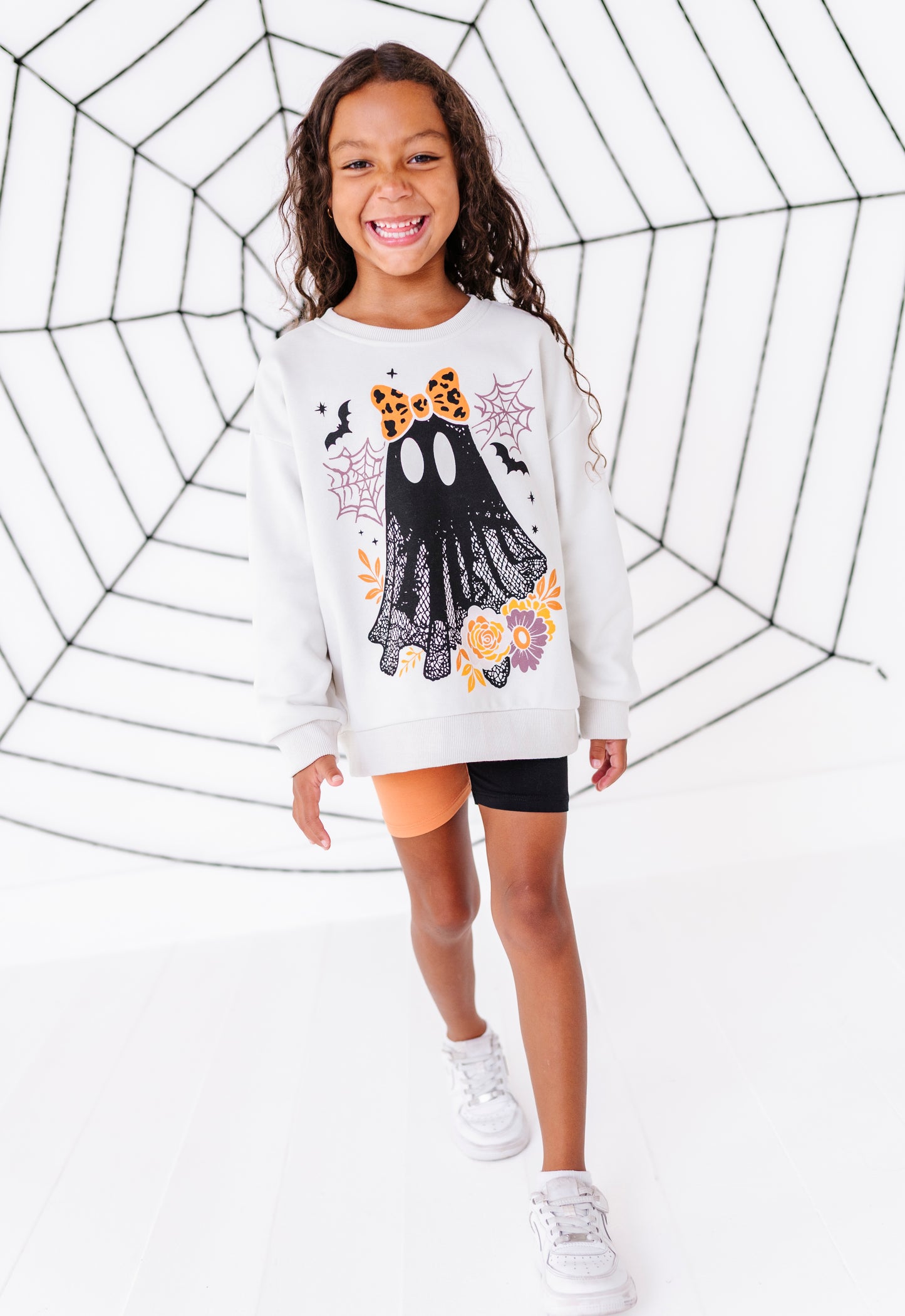 A young girl smiling and walking forward in a white crewneck sweatshirt featuring a black lace ghost graphic with orange and yellow floral accents, paired with black shorts and white sneakers, set against a white studio background with a black spiderweb pattern.