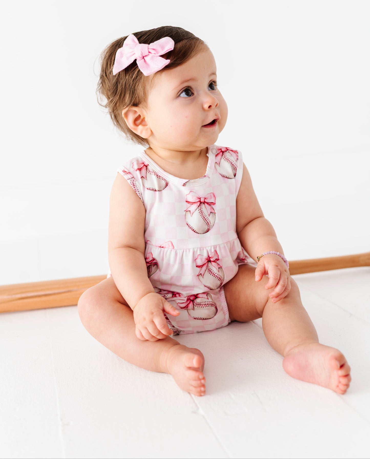 Baby sitting on a white surface wearing a pink baseball romper with bow details.