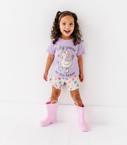 Child wearing a purple shirt with a cartoon goose and silly goose on the loose text, polka dot shorts, and pink rain boots on a white background