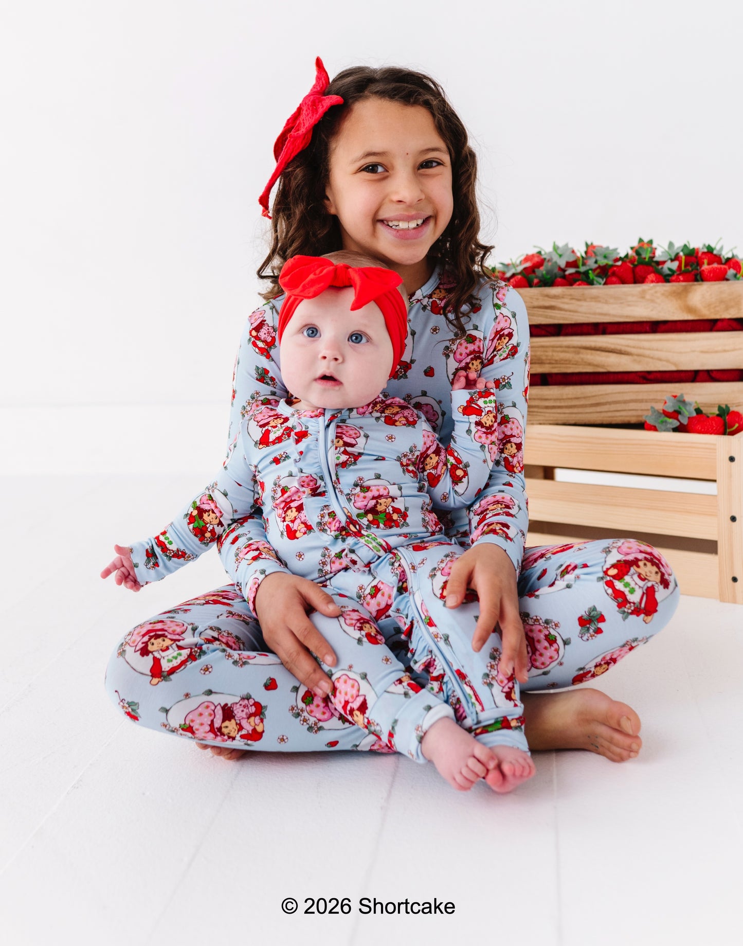 Two children wearing matching Strawberry Shortcake™ pajamas sitting on a white surface with a wooden crate and strawberries in the background.