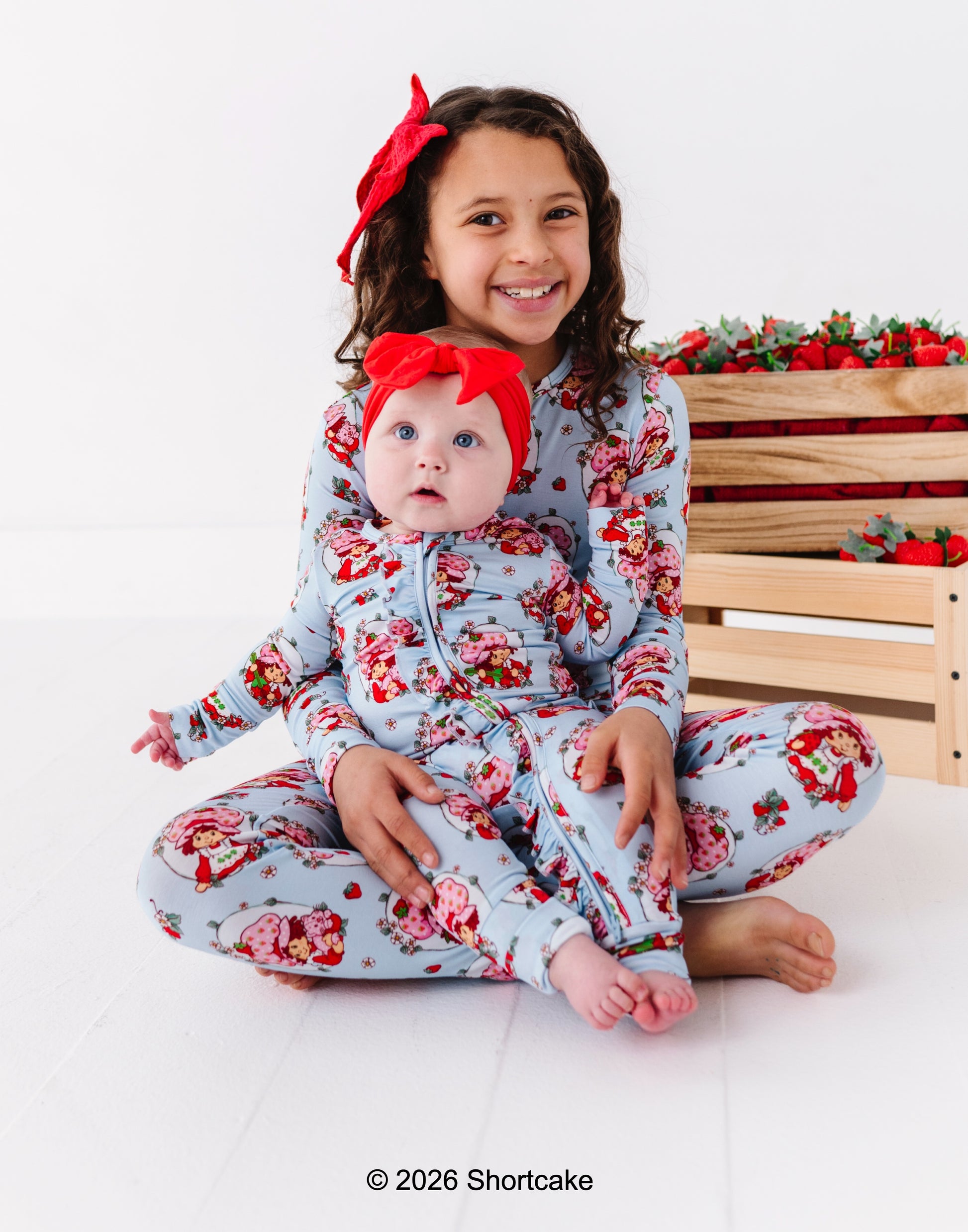 Two children wearing matching Strawberry Shortcake™ pajamas sitting on a white surface with a wooden crate and strawberries in the background.