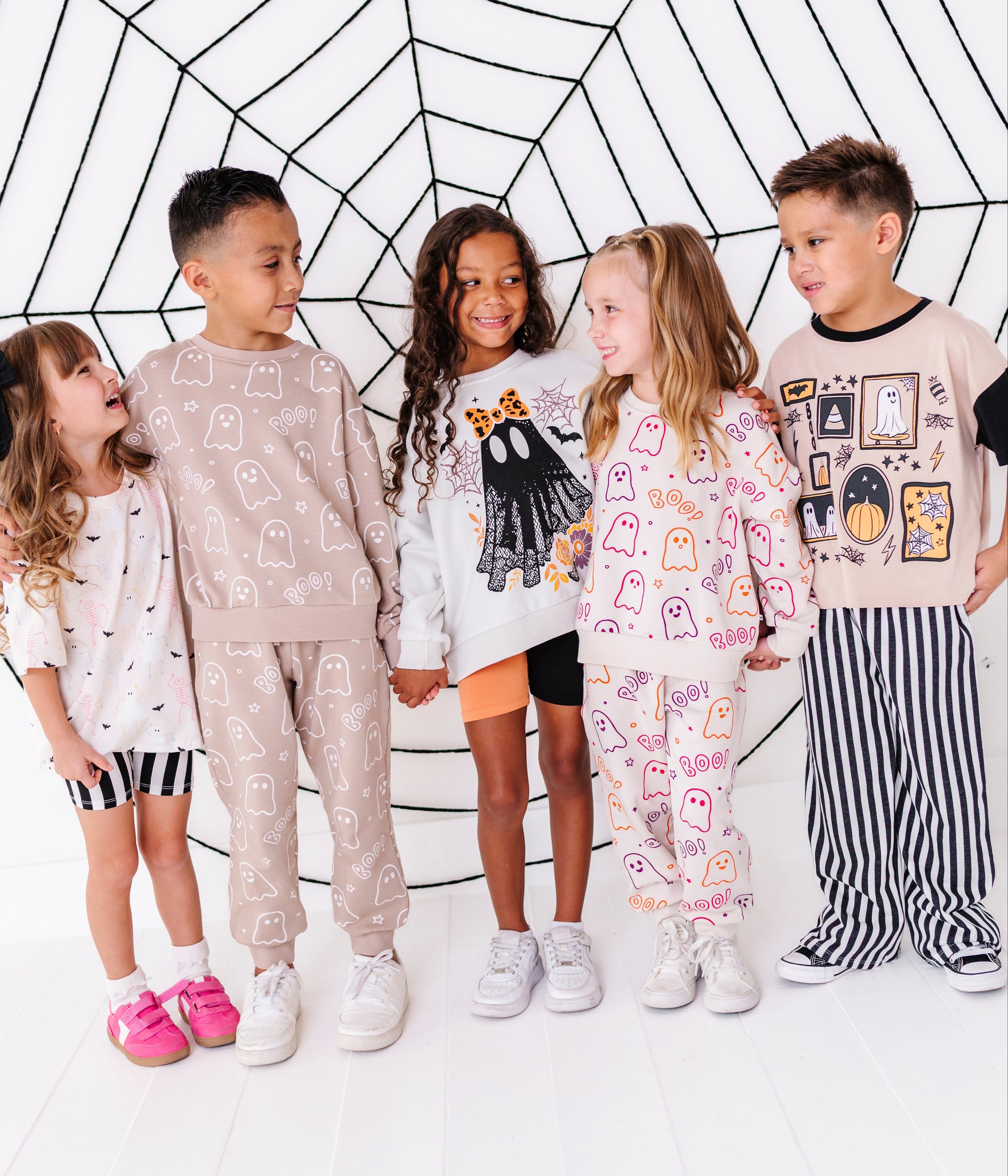 Five children wearing matching Halloween-themed pajama sets stand together in front of a white studio backdrop with a black spiderweb graphic. The pajamas feature ghost and spider motifs in various colors including beige, white, pink, and black.