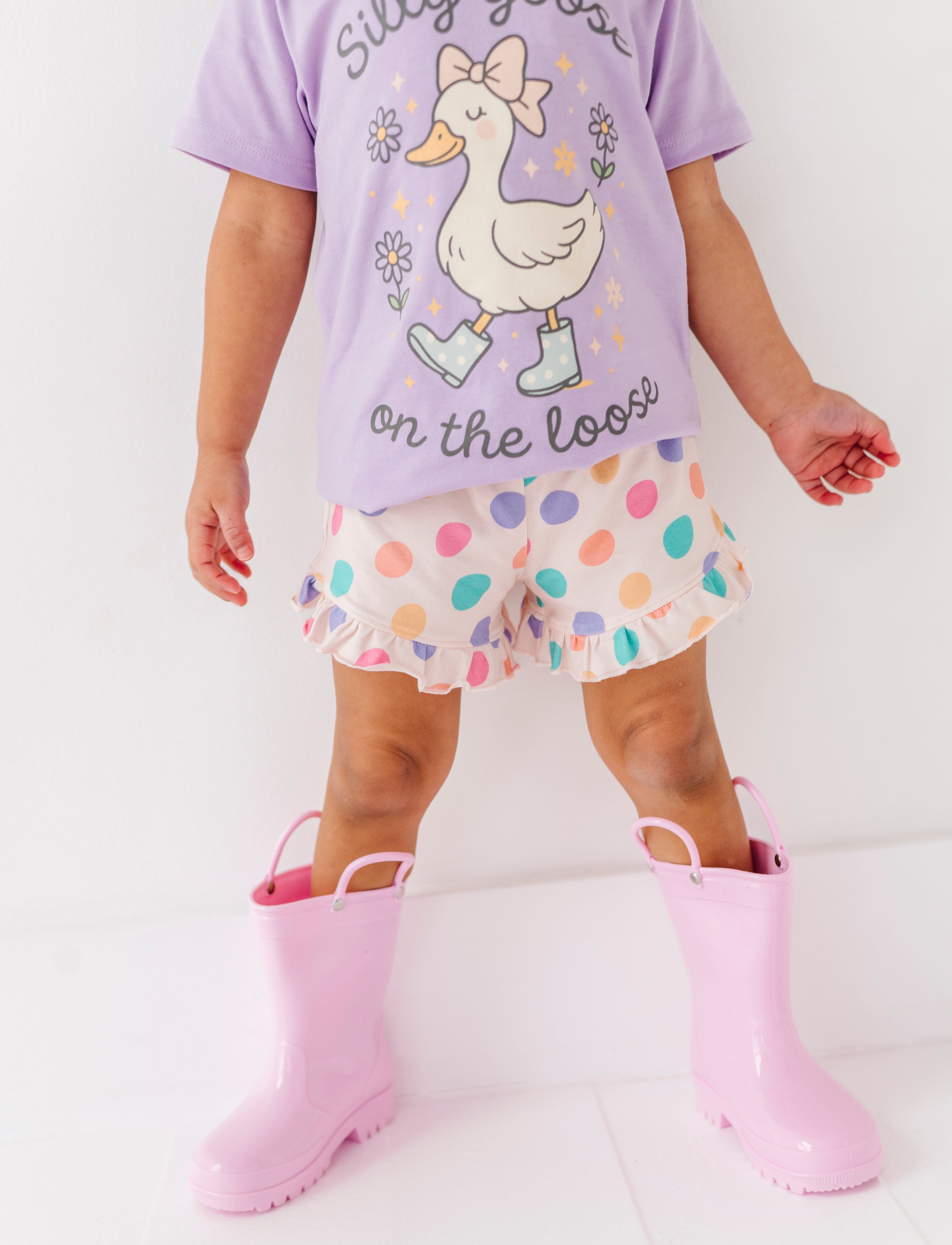 Child wearing a purple shirt with a goose design, colorful bamboo shorts, and pink rain boots on a white background