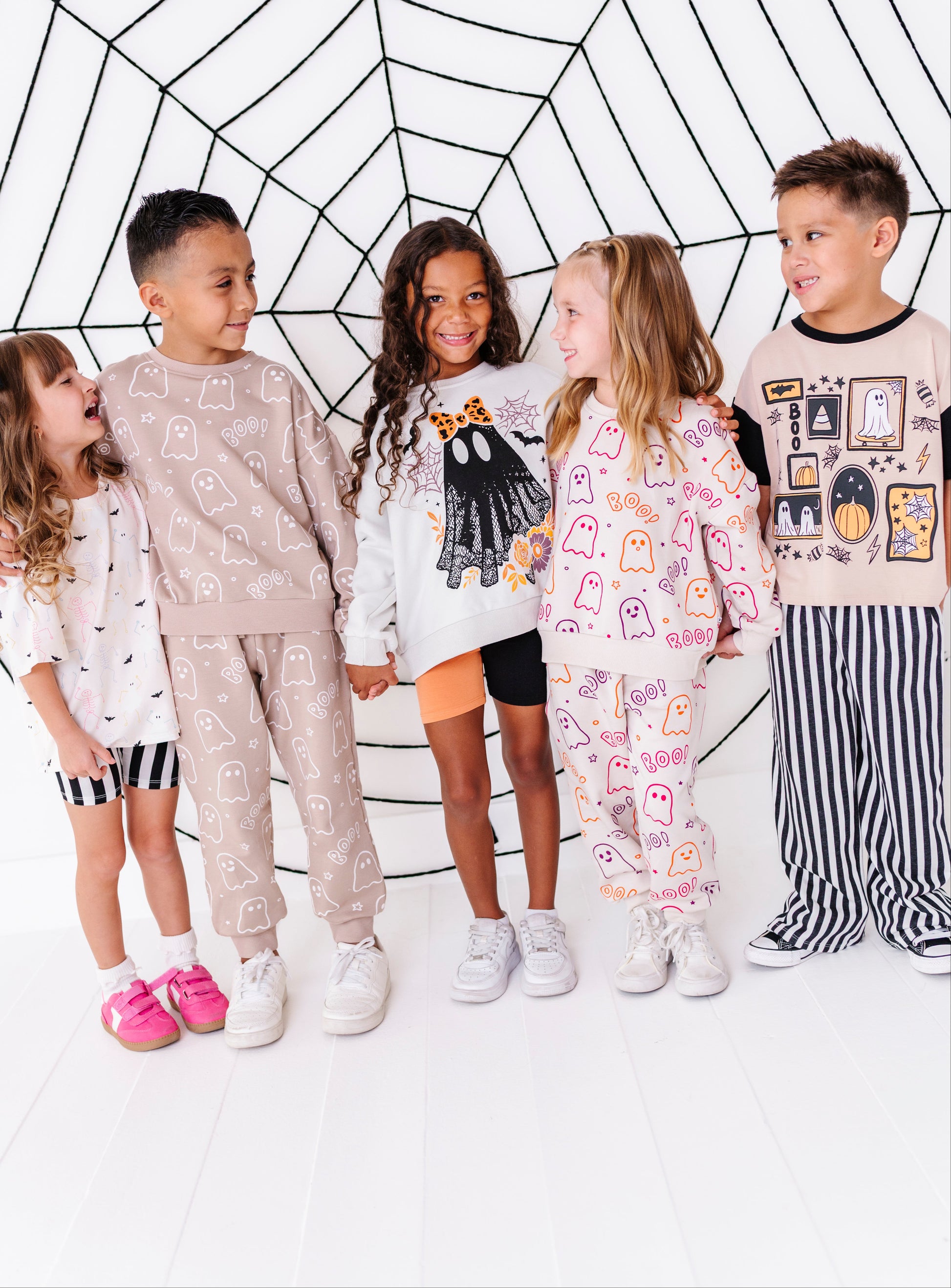 Five children standing together in front of a white wall with a black spiderweb graphic, wearing matching pajama sets with spooky cartoon prints.