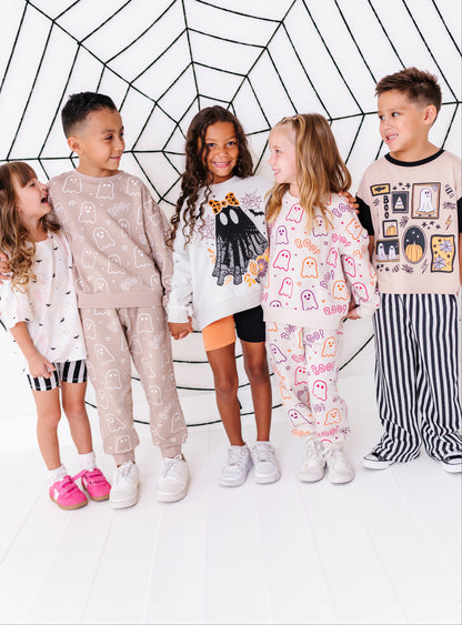Five children standing together in front of a white wall with a black spiderweb graphic, wearing matching pajama sets with spooky cartoon prints.