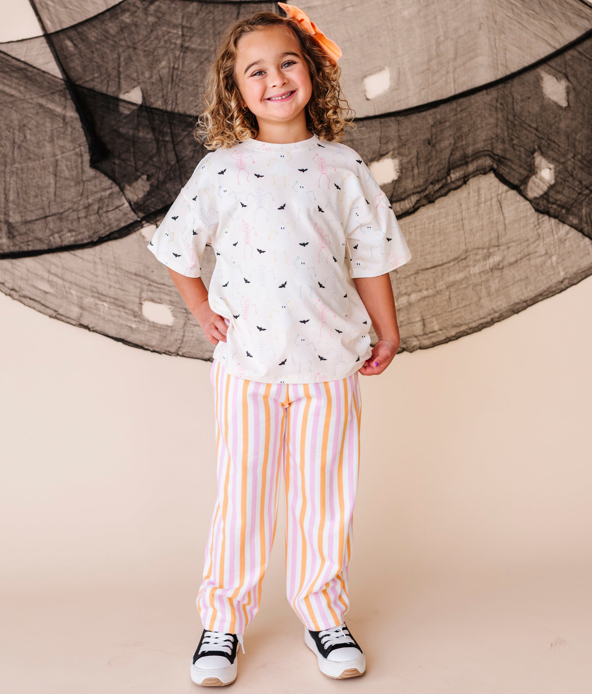 Child wearing a cream shirt with halloween patterns and striped pants standing in front of large netting.