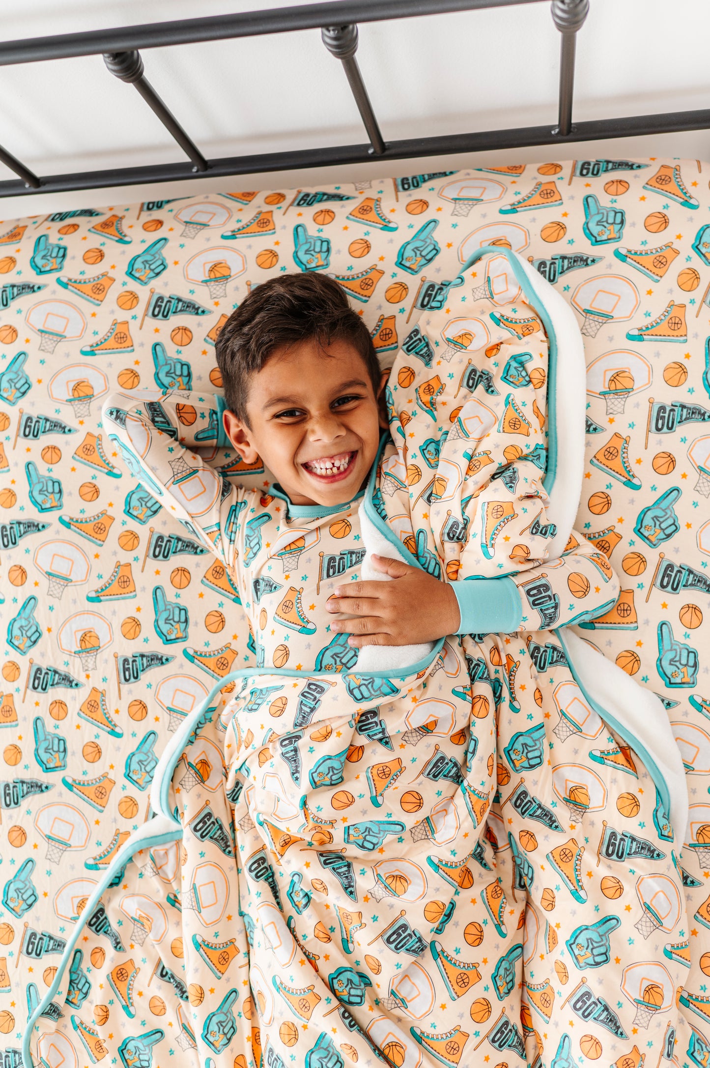 Child wearing sports themed bamboo pajamas, wrapped in a basketball themed blanket, lying on a sports focused bamboo sheet