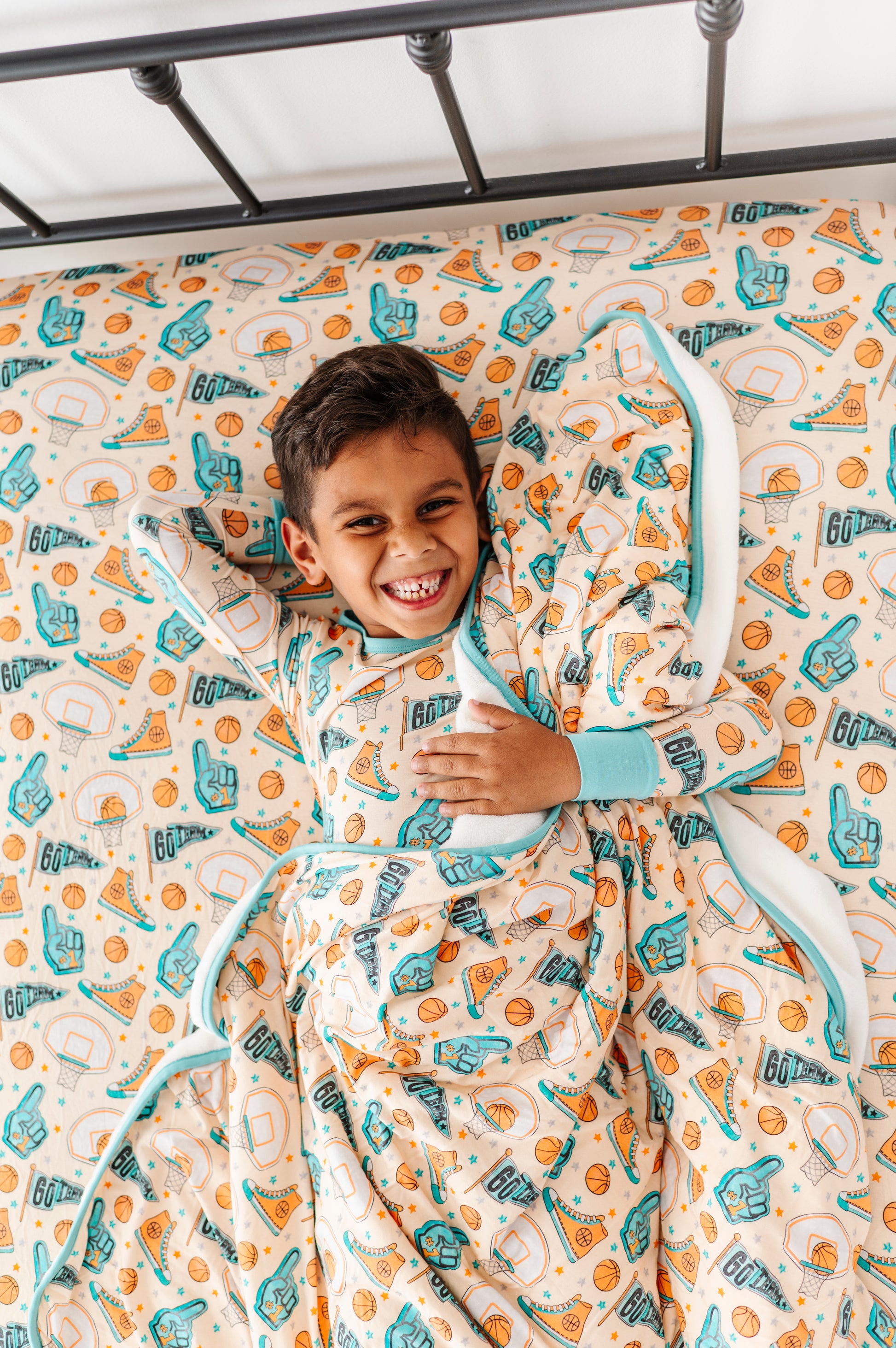 Child wearing sports themed bamboo pajamas, wrapped in a basketball themed blanket, lying on a sports focused bamboo sheet