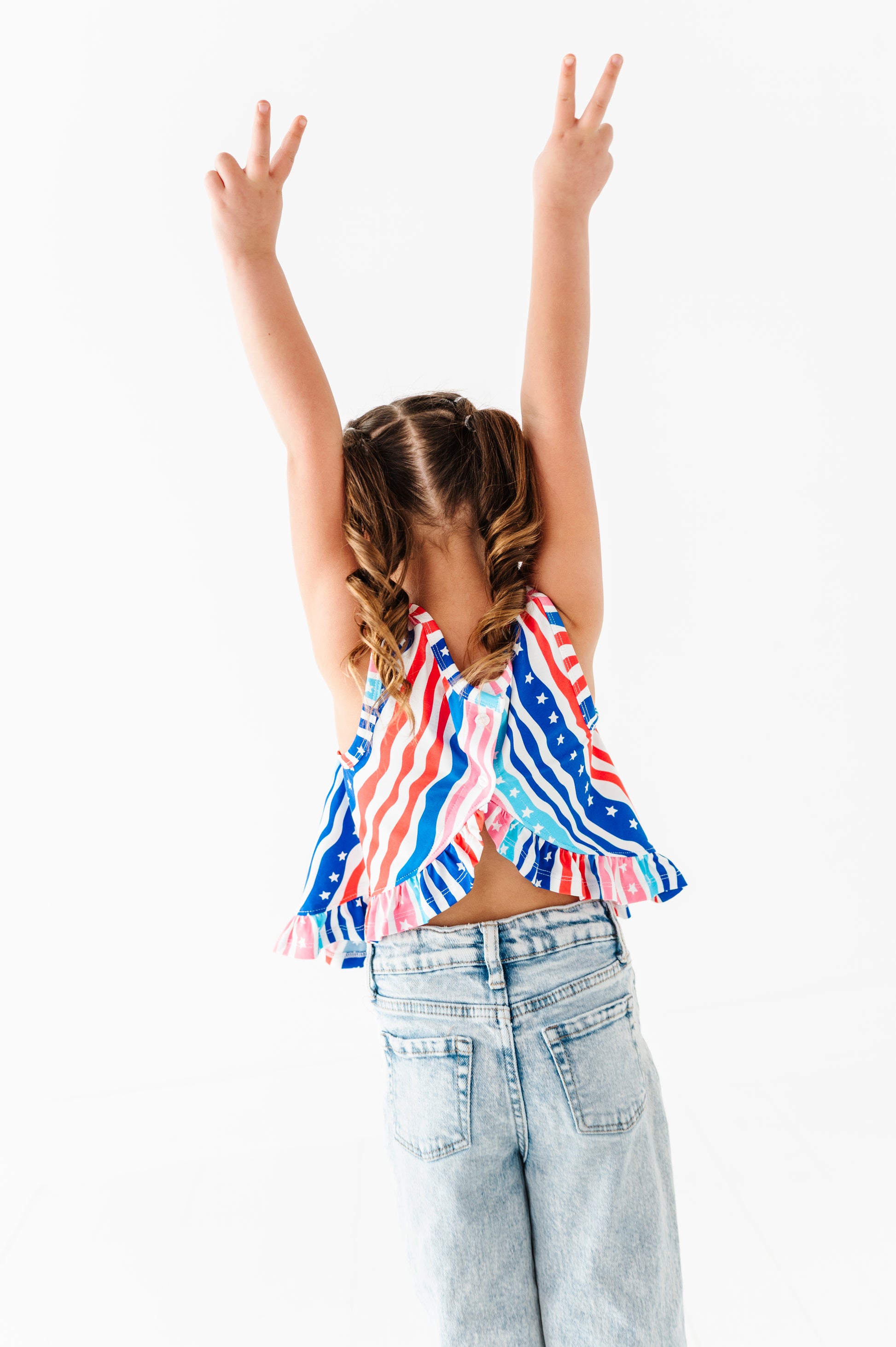 Young girl wearing a colorful striped bamboo-cotton top and light blue jeans with arms raised on a white background