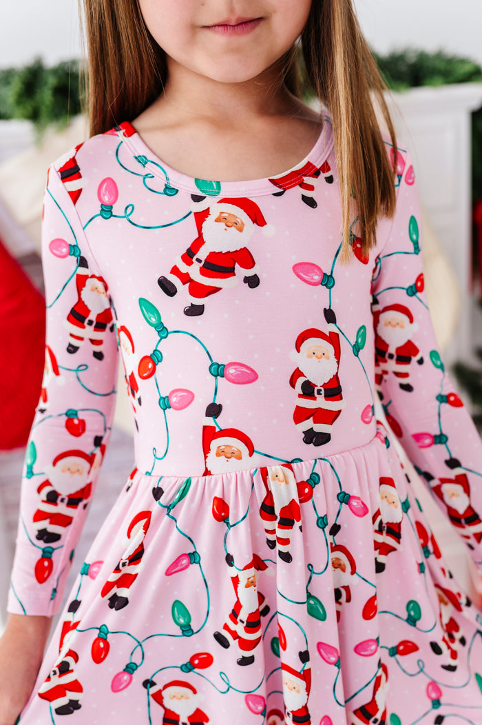 A young girl wearing a pink long-sleeve bamboo dress with a festive Christmas print of Santa Claus and colorful holiday lights.