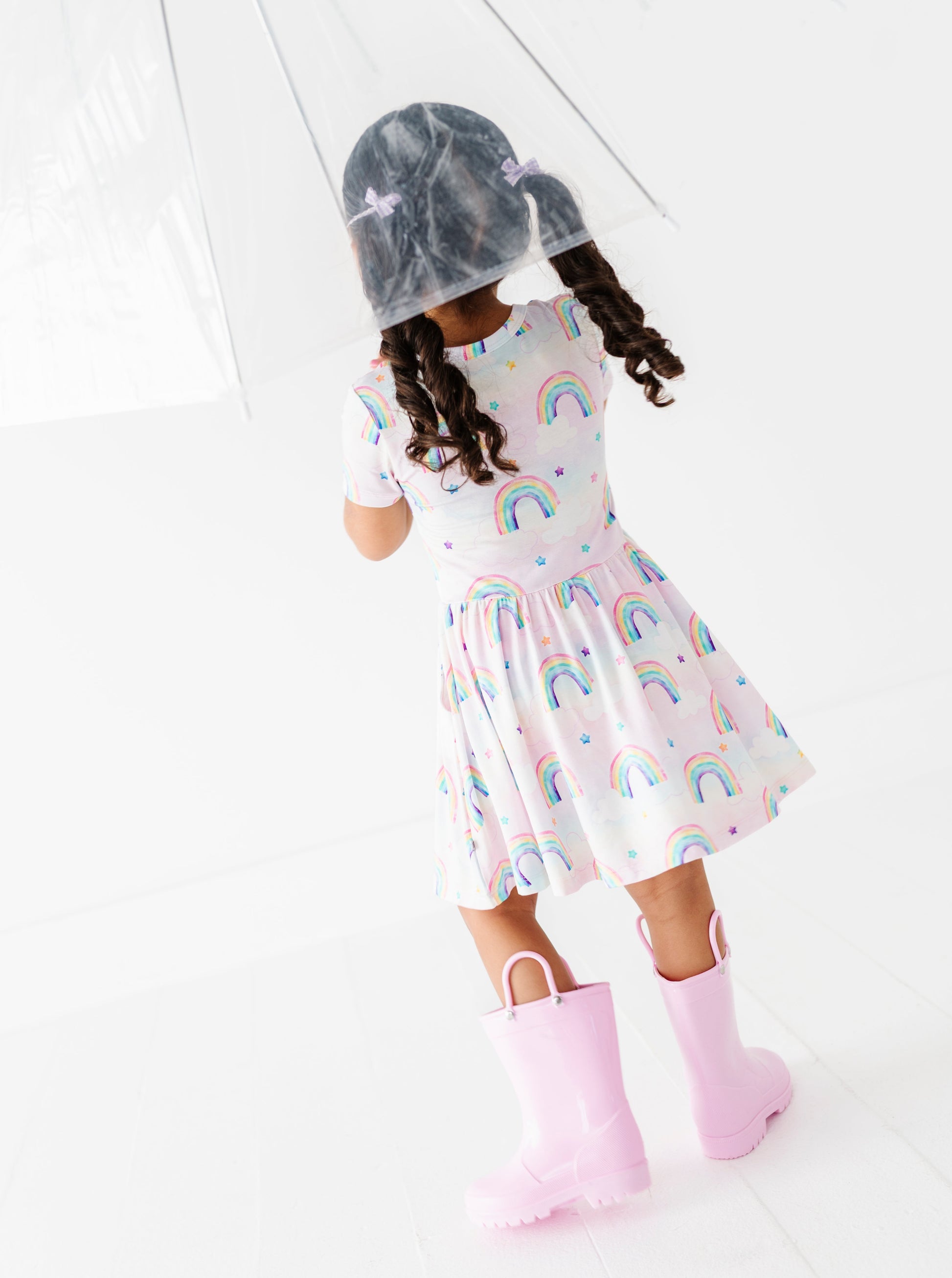 Child wearing a bamboo dress with watercolor rainbow patterns and pink rain boots on a white floor.