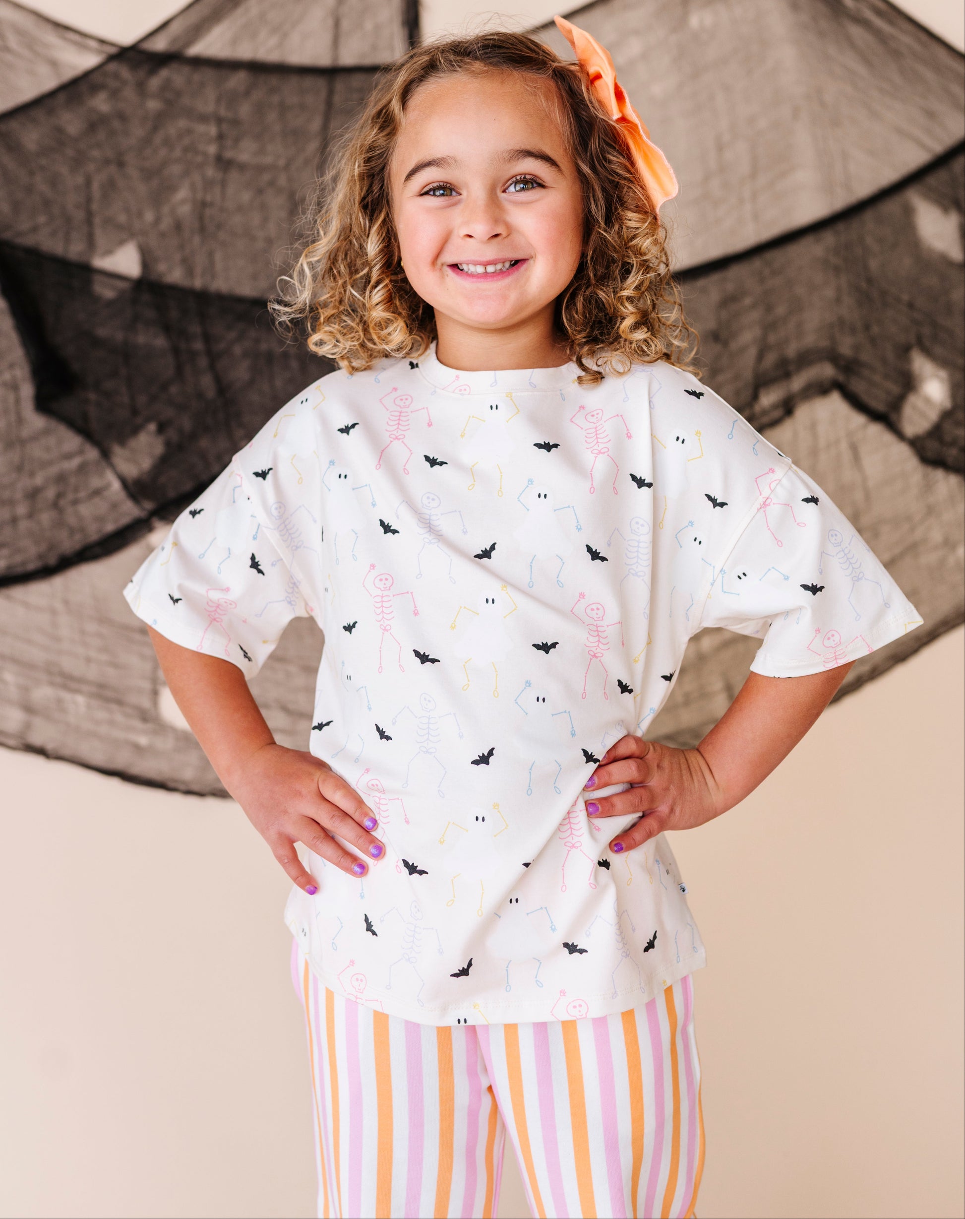 Young girl wearing a cream shirt with halloween designs and striped pants, standing in front of a net.