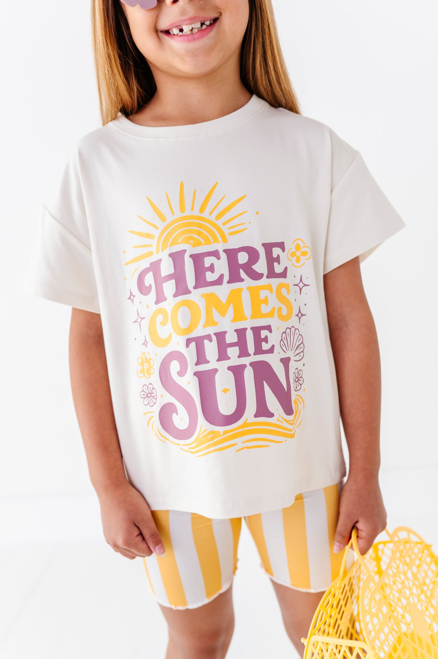 Child wearing a white t-shirt with 'Here Comes the Sun' text and yellow shorts on a white background