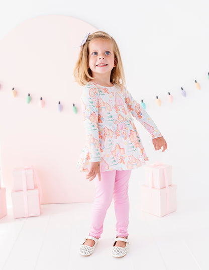 A young girl with blonde hair wearing a pastel pink patterned long-sleeve bamboo peplum top, pink leggings, and white shoes, standing in a bright white studio decorated with pastel pink arches, gift boxes, and a string of colorful lights.