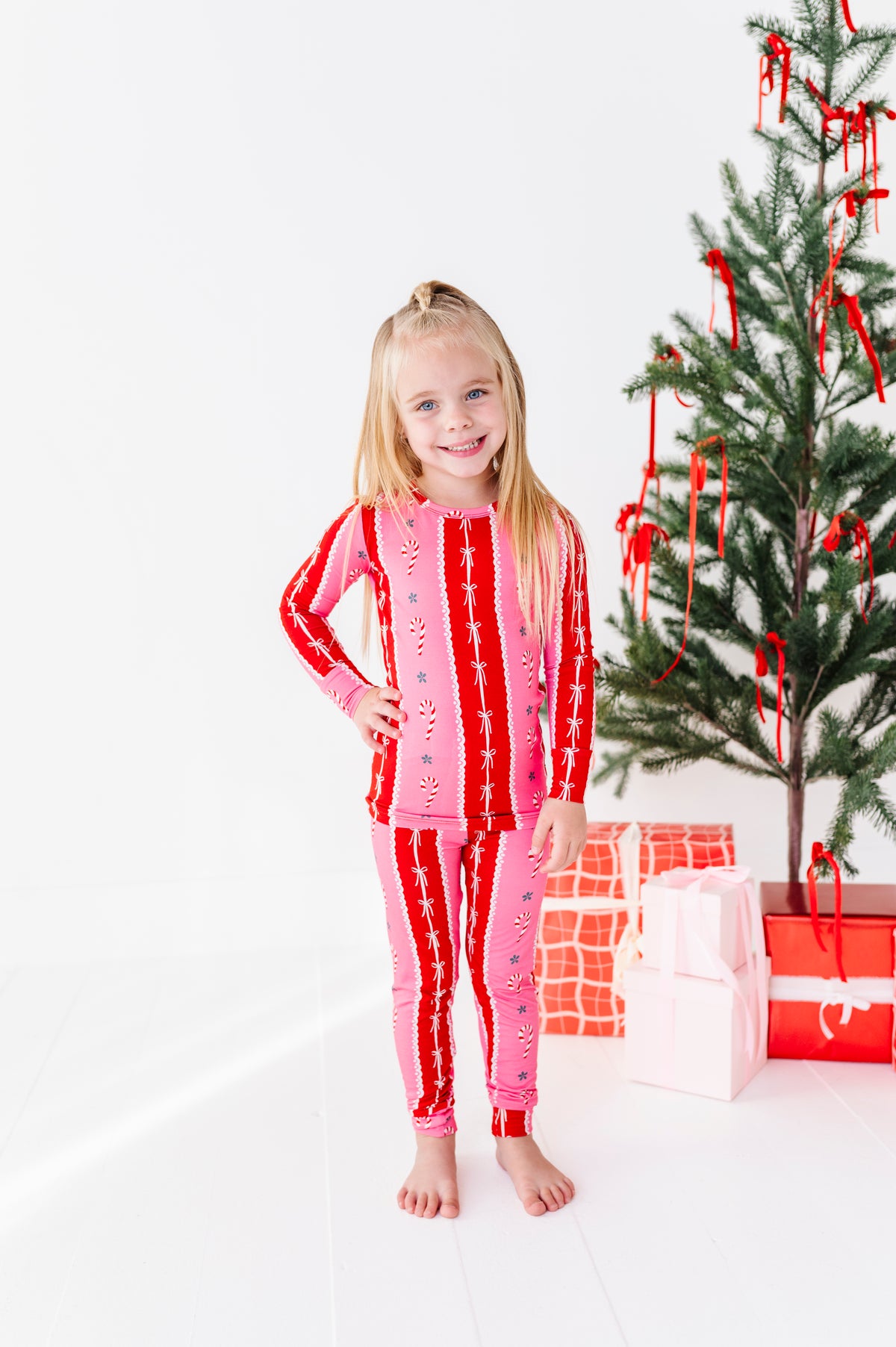 A young girl stands barefoot in a white room wearing a pink and red holiday-themed long-sleeve bamboo pajama set with vertical candy cane stripes. To her right is a decorated Christmas tree and wrapped gift boxes.