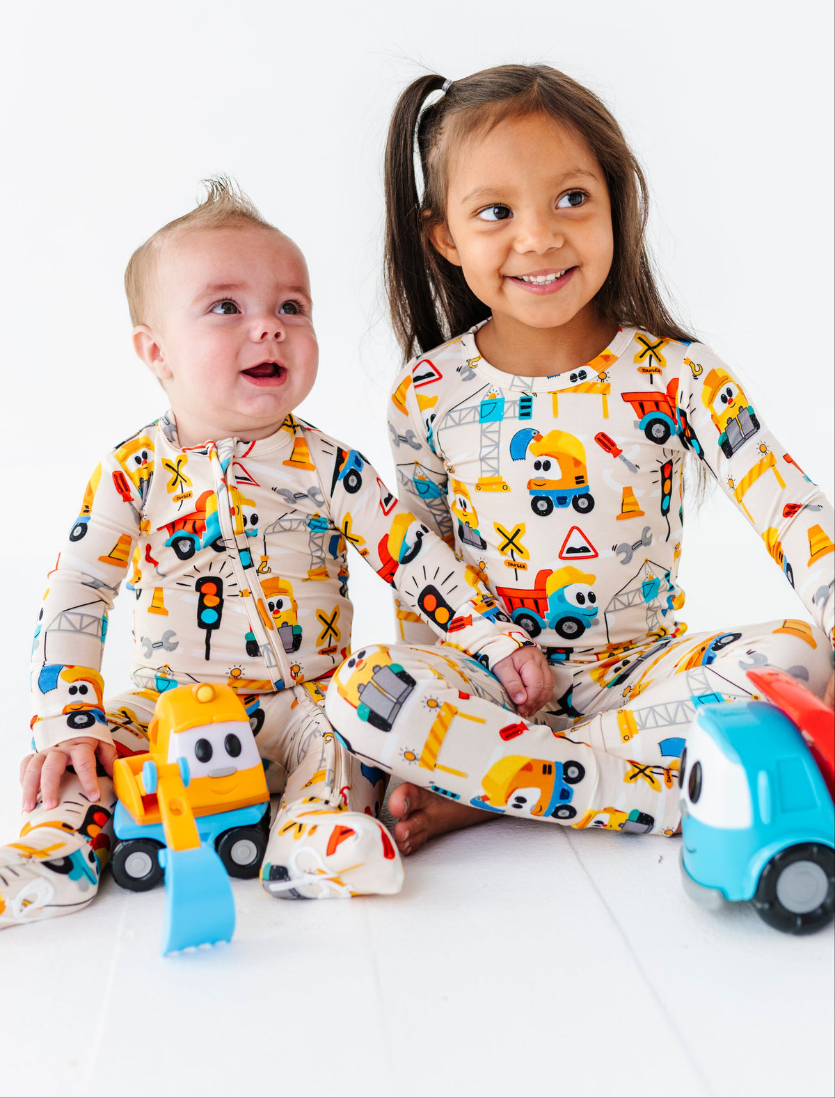 Two children sitting on a white floor wearing matching construction-themed pajamas, with toy vehicles in front of them against a clean white background.