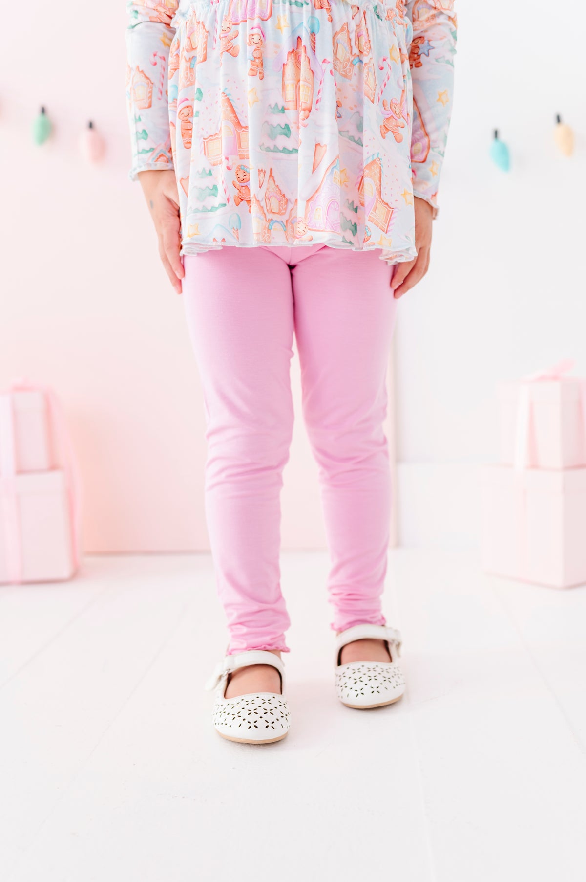 A child wearing pink bamboo leggings, a pastel multicolor patterned long-sleeve peplum top, and white embellished Mary Jane shoes, standing against a soft pink studio background.