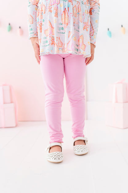 A child wearing pink bamboo leggings, a pastel multicolor patterned long-sleeve peplum top, and white embellished Mary Jane shoes, standing against a soft pink studio background.