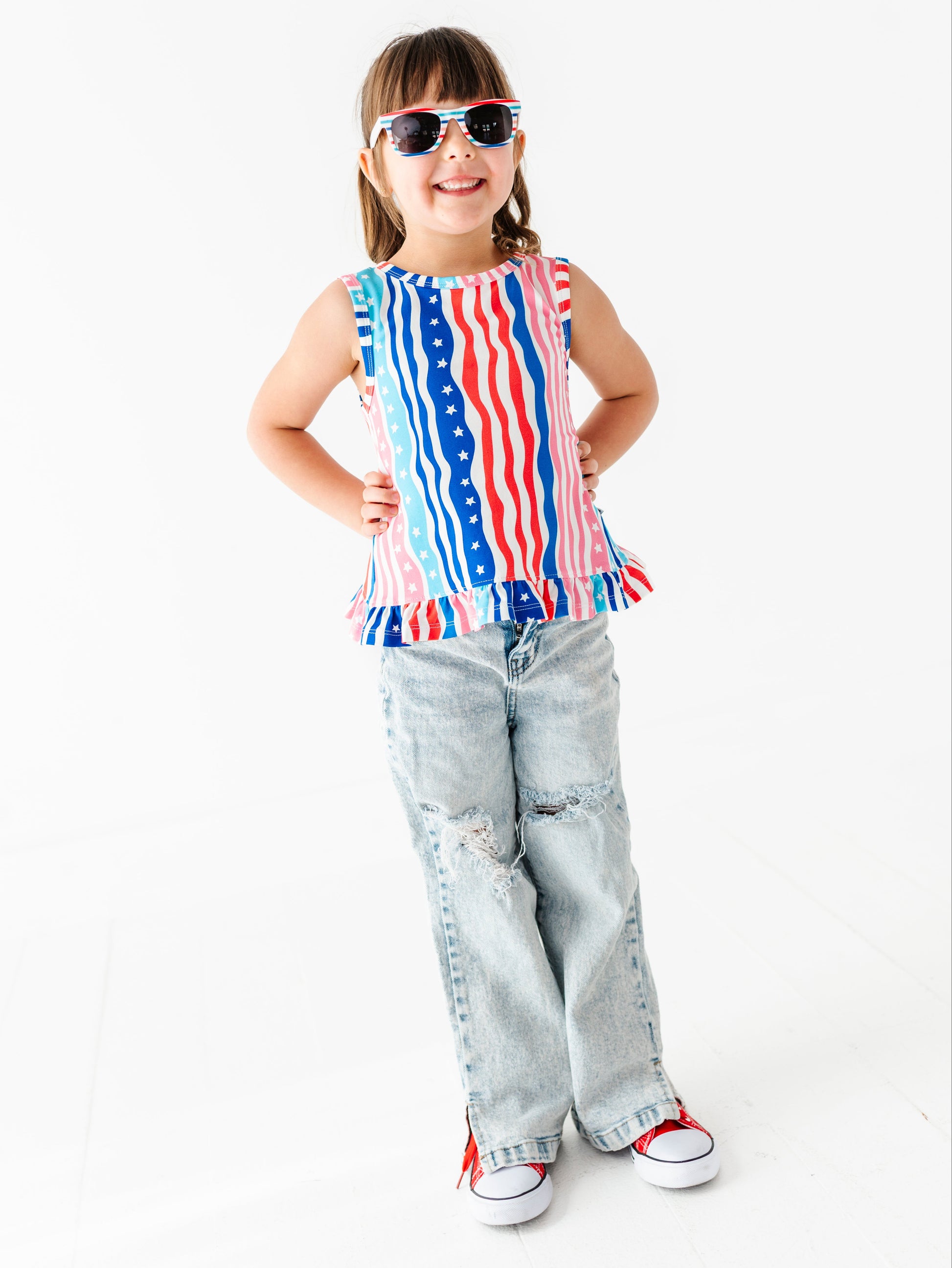 Child wearing a sleeveless bamboo-cotton swing top with an American flag design and ripped jeans on a white background