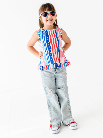 Child wearing a sleeveless bamboo-cotton swing top with an American flag design and ripped jeans on a white background