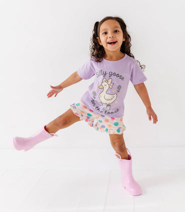 Child wearing a purple shirt with a goose design, colorful bamboo shorts, and pink rain boots on a white background