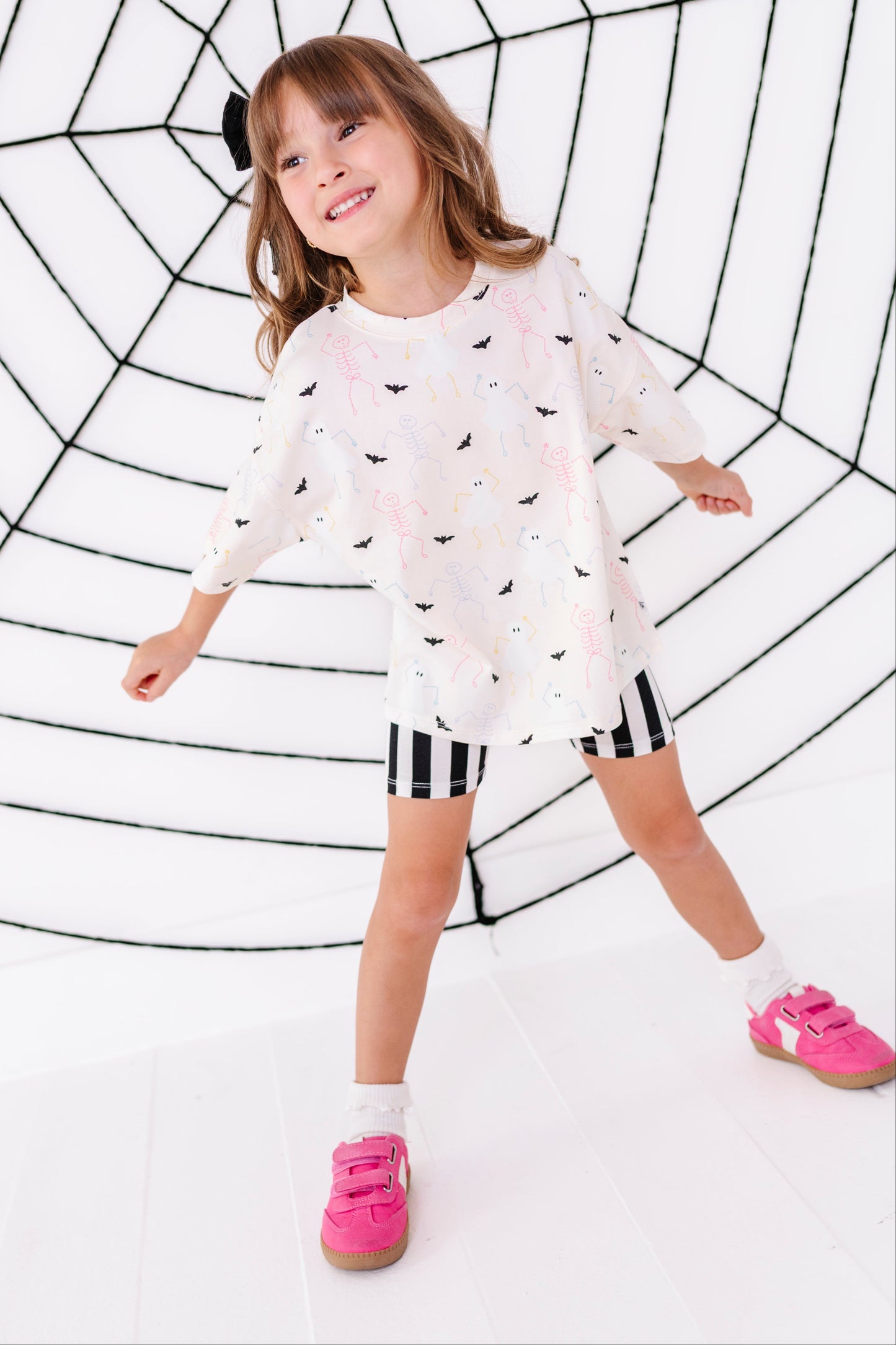 A young girl with brown hair wearing a white long-sleeve t-shirt with a black bat pattern, black and white striped shorts, and pink sneakers, posing in front of a white studio backdrop with a black spiderweb graphic.
