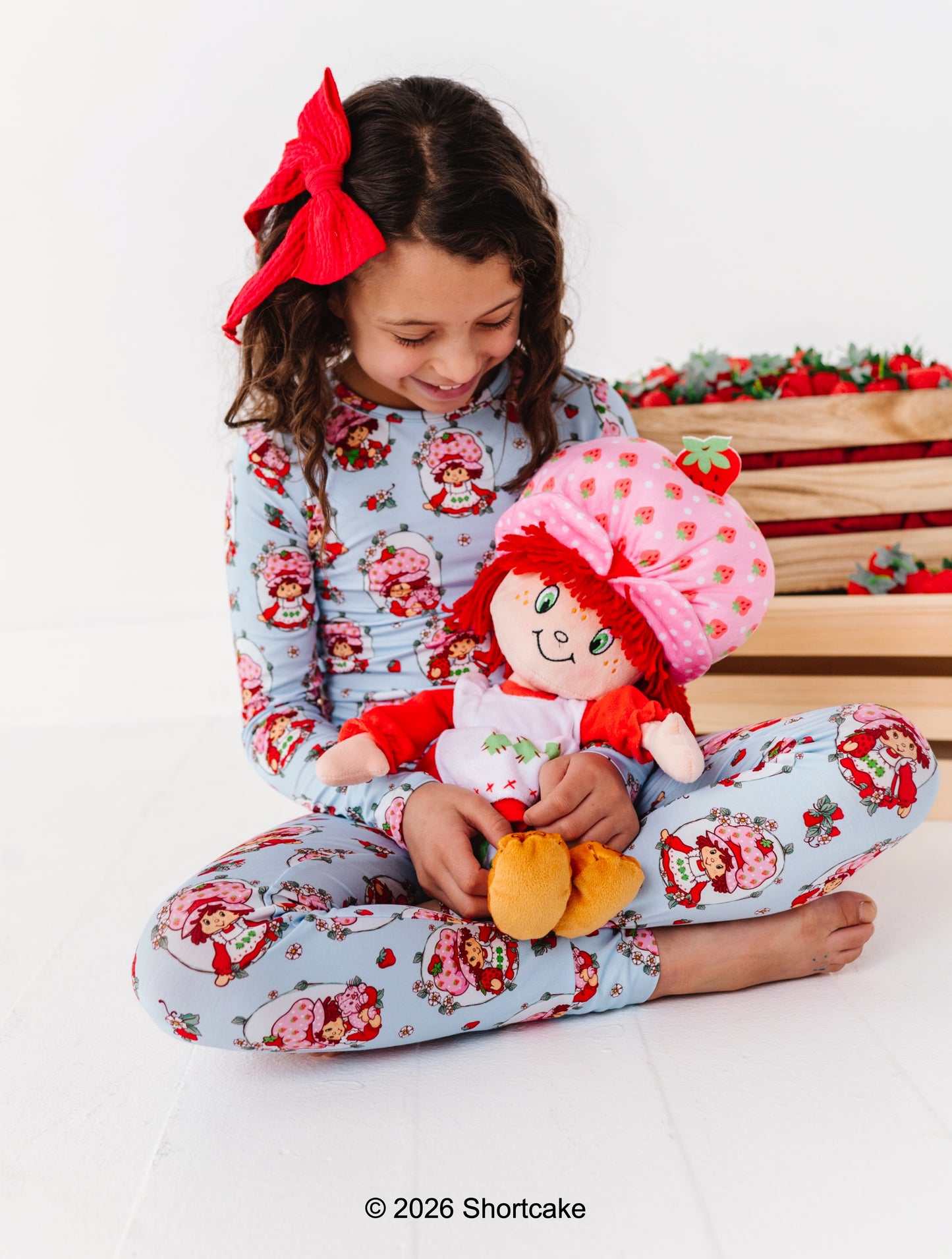Little girl wearing Strawberry Shortcake™ bamboo pajamas, holding a Strawberry Shortcake™ doll against a white background 