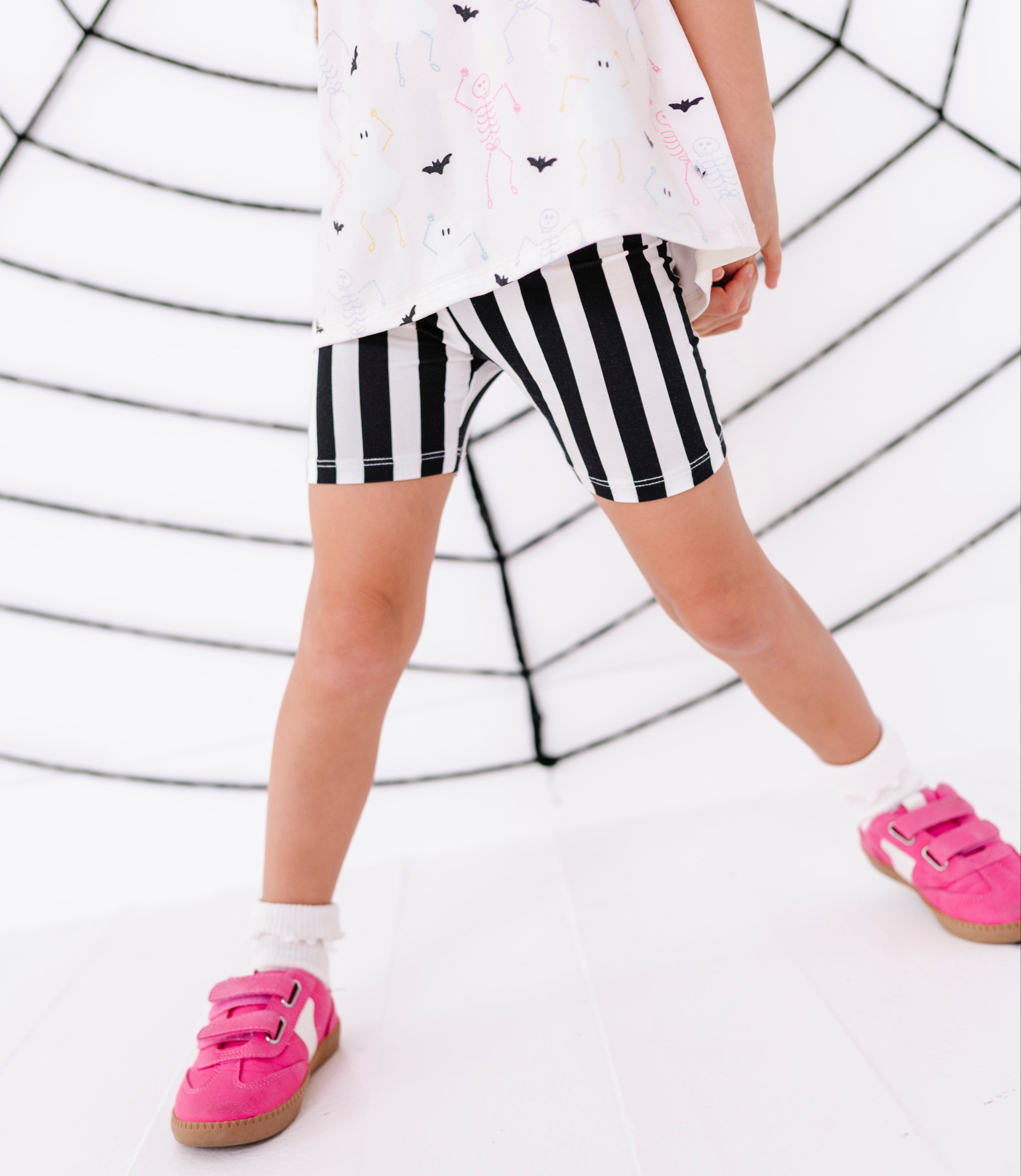 A child wearing black and white vertically striped shorts, a white t-shirt with a black bat pattern, white socks, and bright pink sneakers, standing in front of a white background with a black spiderweb graphic.