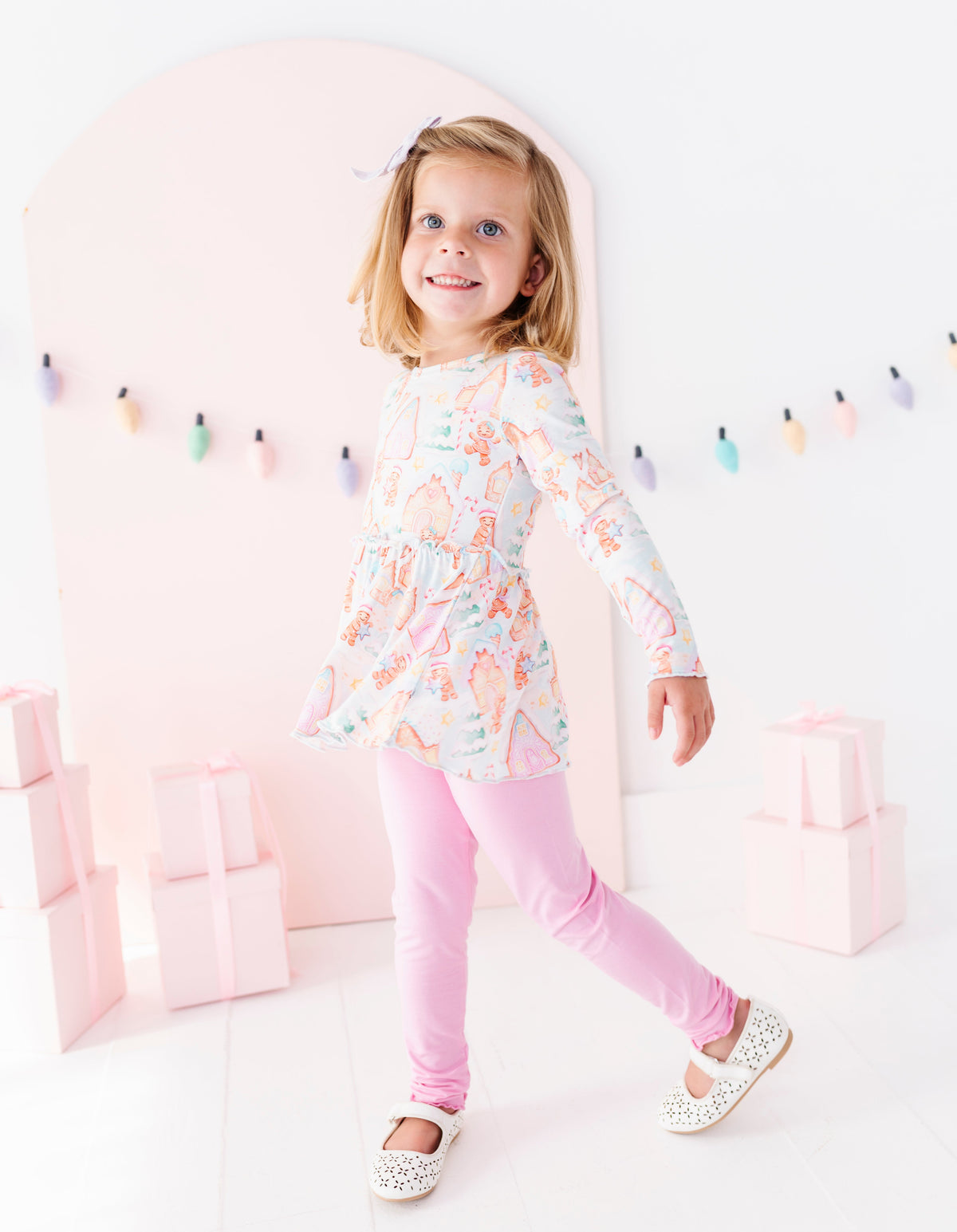 A young girl with blonde hair wearing a pastel multicolor ice cream print tunic top and pink leggings, posing in a bright, whimsical studio setting with pink arches and gift boxes.