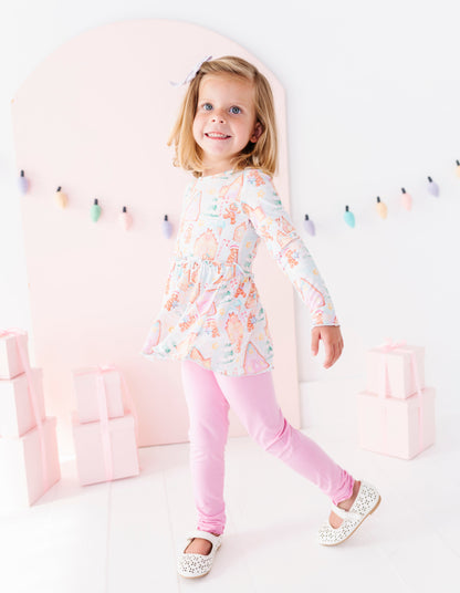 A young girl with blonde hair wearing a pastel multicolor ice cream print tunic top and pink leggings, posing in a bright, whimsical studio setting with pink arches and gift boxes.