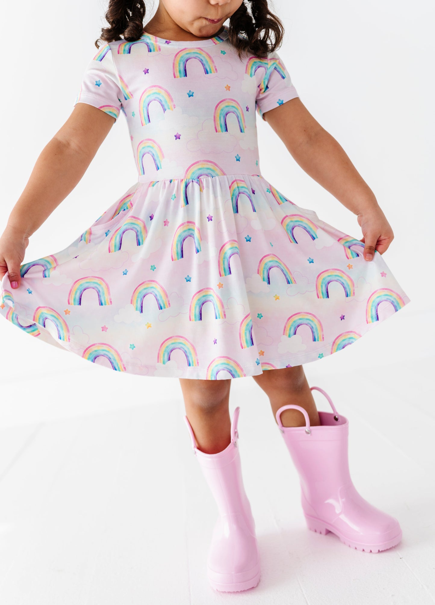 Child wearing a bamboo dress with watercolor rainbow pattern and pink rain boots on a white background