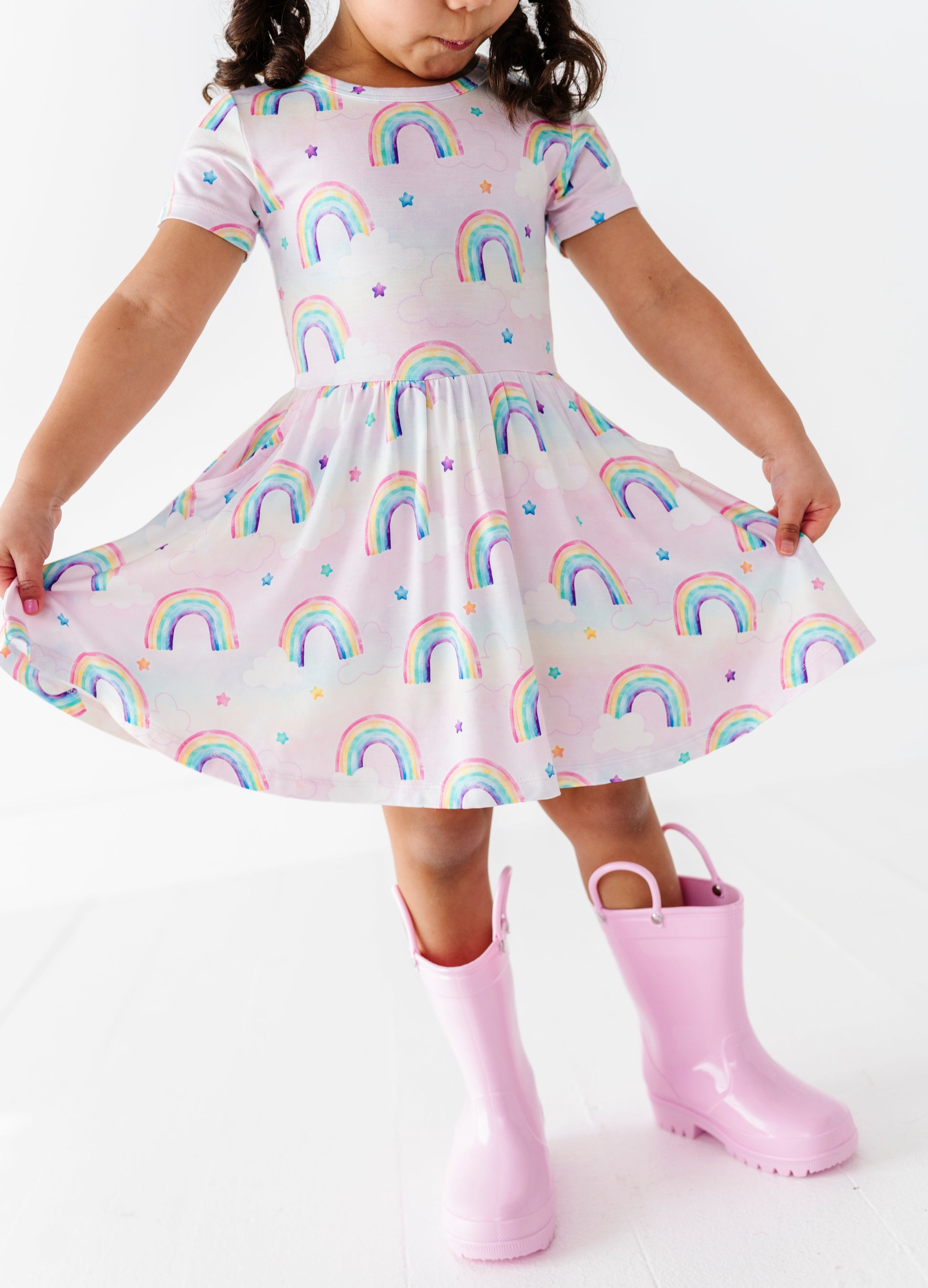 Child wearing a bamboo dress with watercolor rainbow pattern and pink rain boots on a white background
