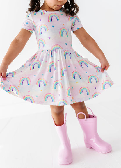 Child wearing a bamboo dress with watercolor rainbow pattern and pink rain boots on a white background
