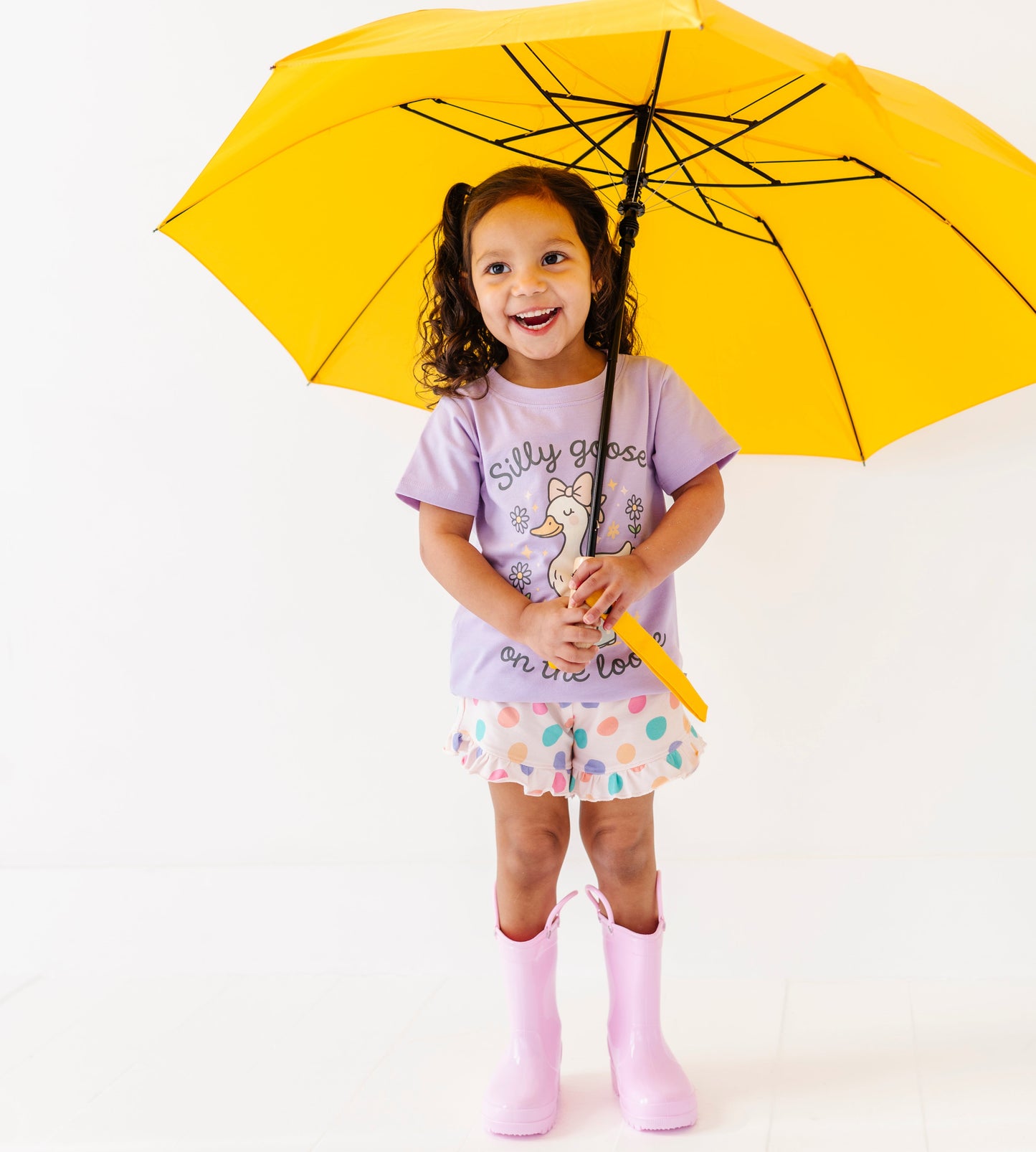Child wearing a purple shirt with a cartoon goose and silly goose on the loose text, polka dot shorts, and pink rain boots on a white background