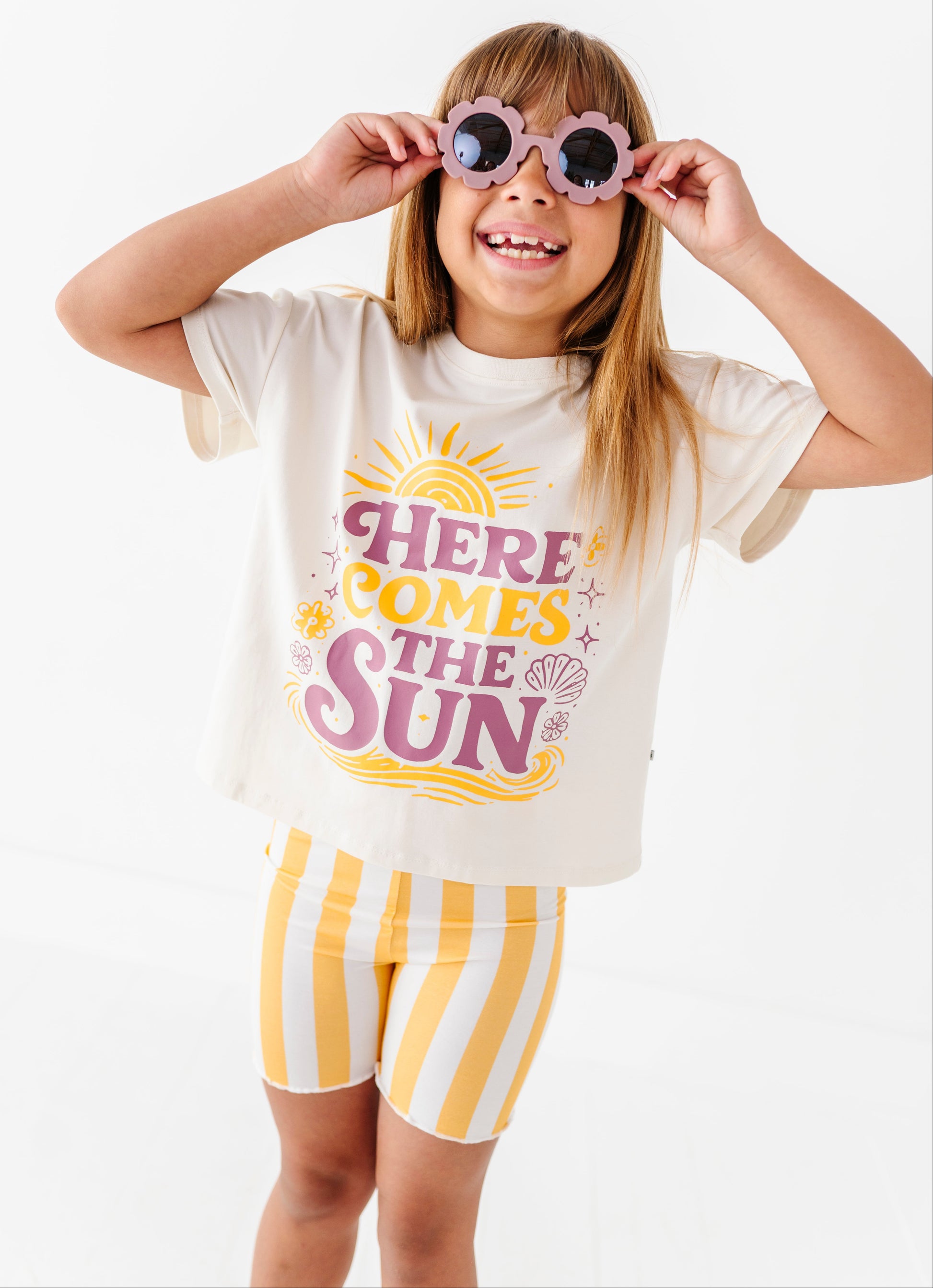 Child wearing a 'Here Comes the Sun' t-shirt and yellow striped shorts on a white background