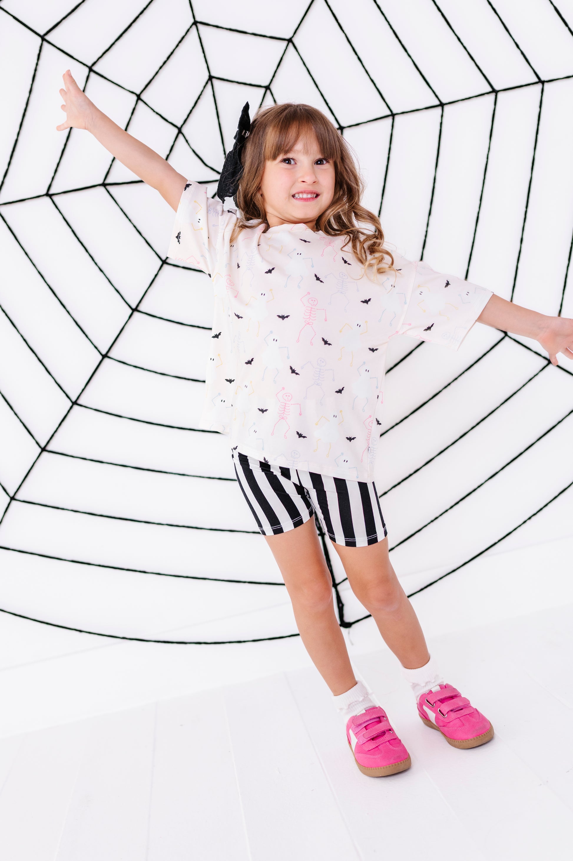 A young girl posing in a white t-shirt with a black bat pattern, black and white striped shorts, and bright pink shoes, set against a white background with a large black spiderweb graphic.