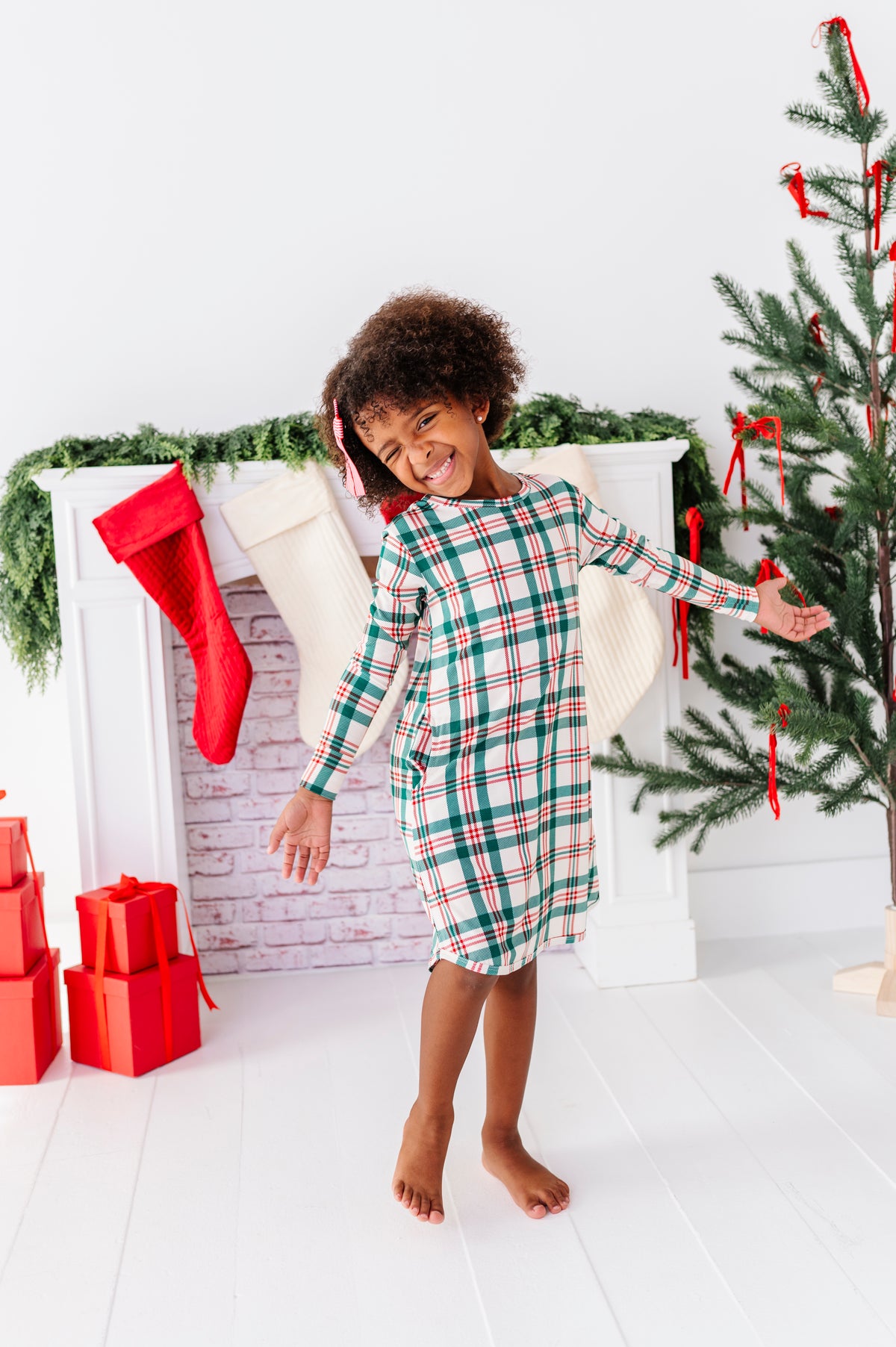 A little girl wearing a bamboo gown in a classic Christmas plaid print, standing in front of a fireplace with stockings and a decorated Christmas tree