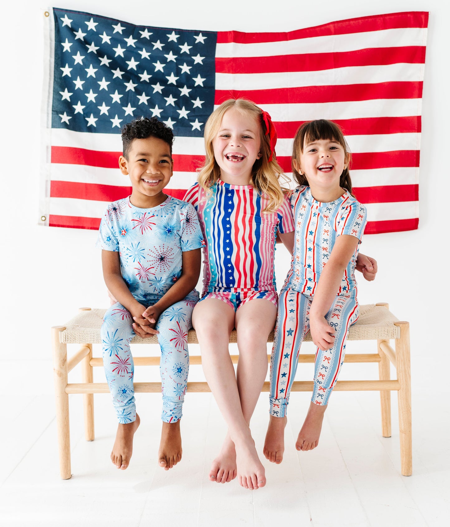 Three children in bamboo pajamas sitting on a bench with an American flag in the background