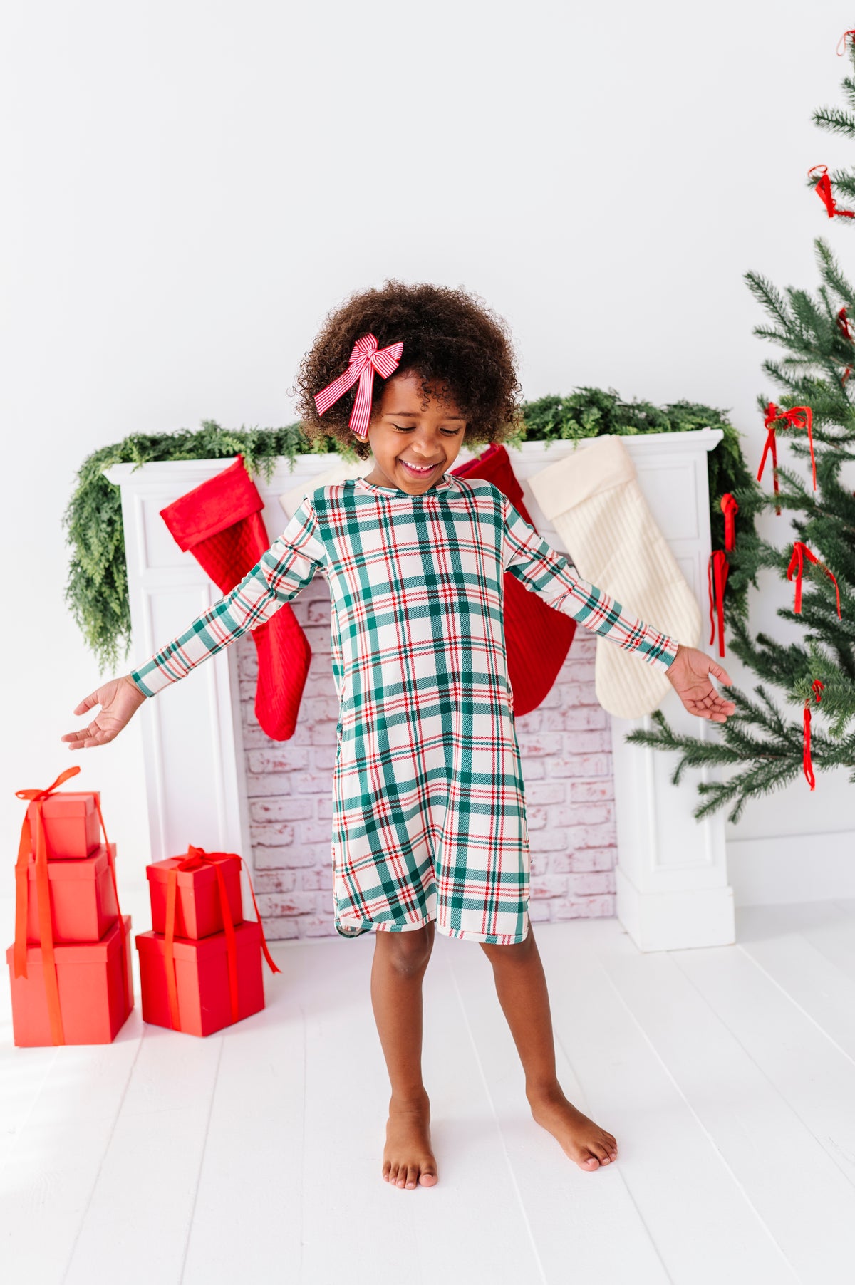 A little girl wearing a bamboo nightgown in a classic Christmas plaid print, standing in front of a fireplace with stockings 