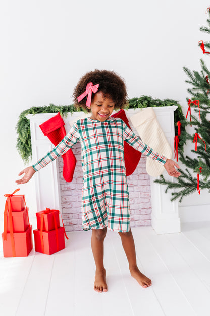 A little girl wearing a bamboo nightgown in a classic Christmas plaid print, standing in front of a fireplace with stockings 