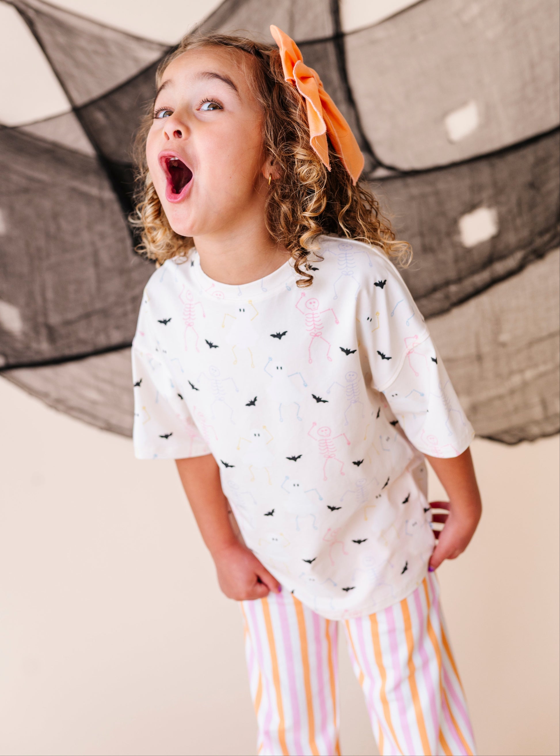Child wearing a cream shirt with halloween patterns and striped pants, standing in front of a black net.